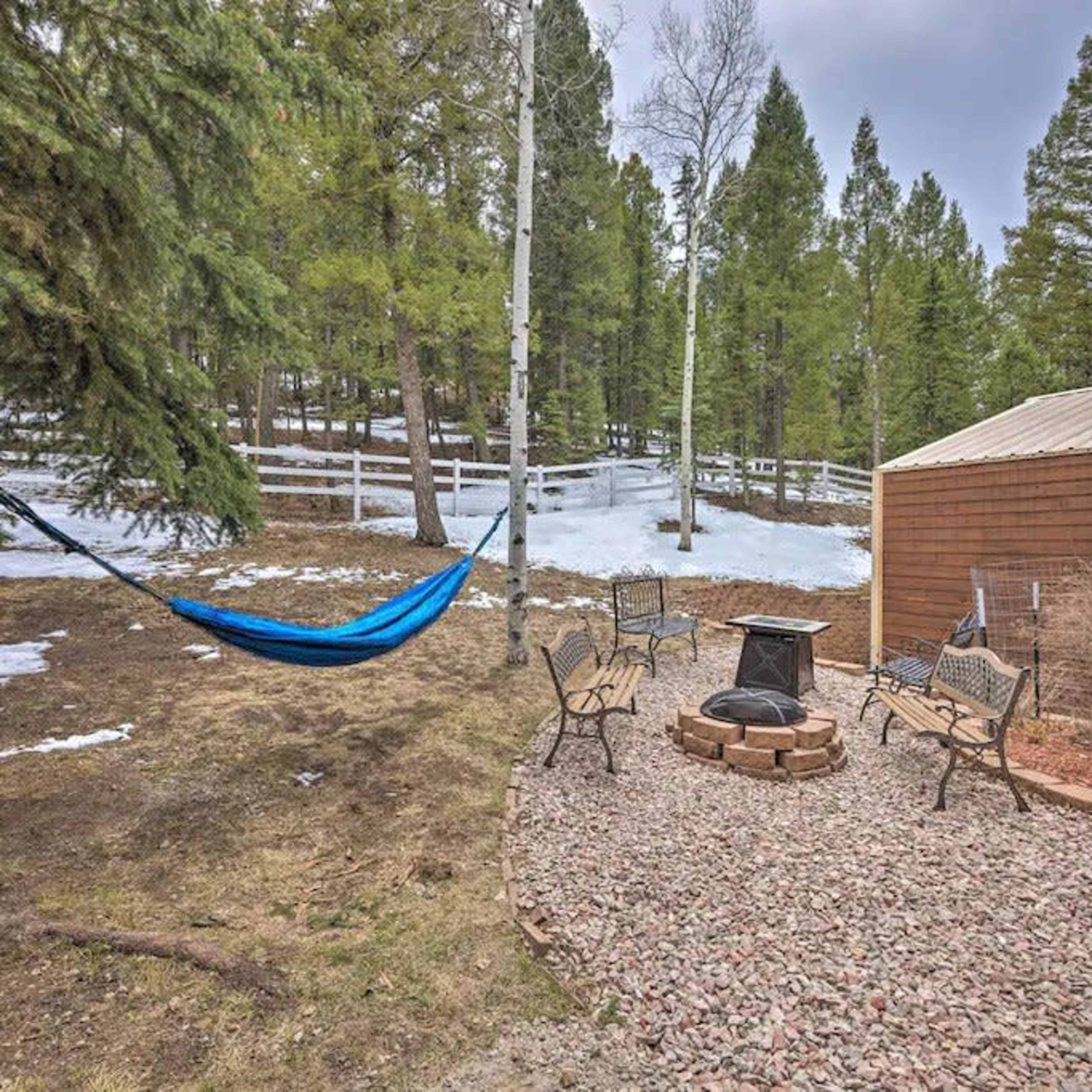 A blue hammock is suspended between two trees near a stone fire pit surrounded by seating in a snow-covered wooded area.