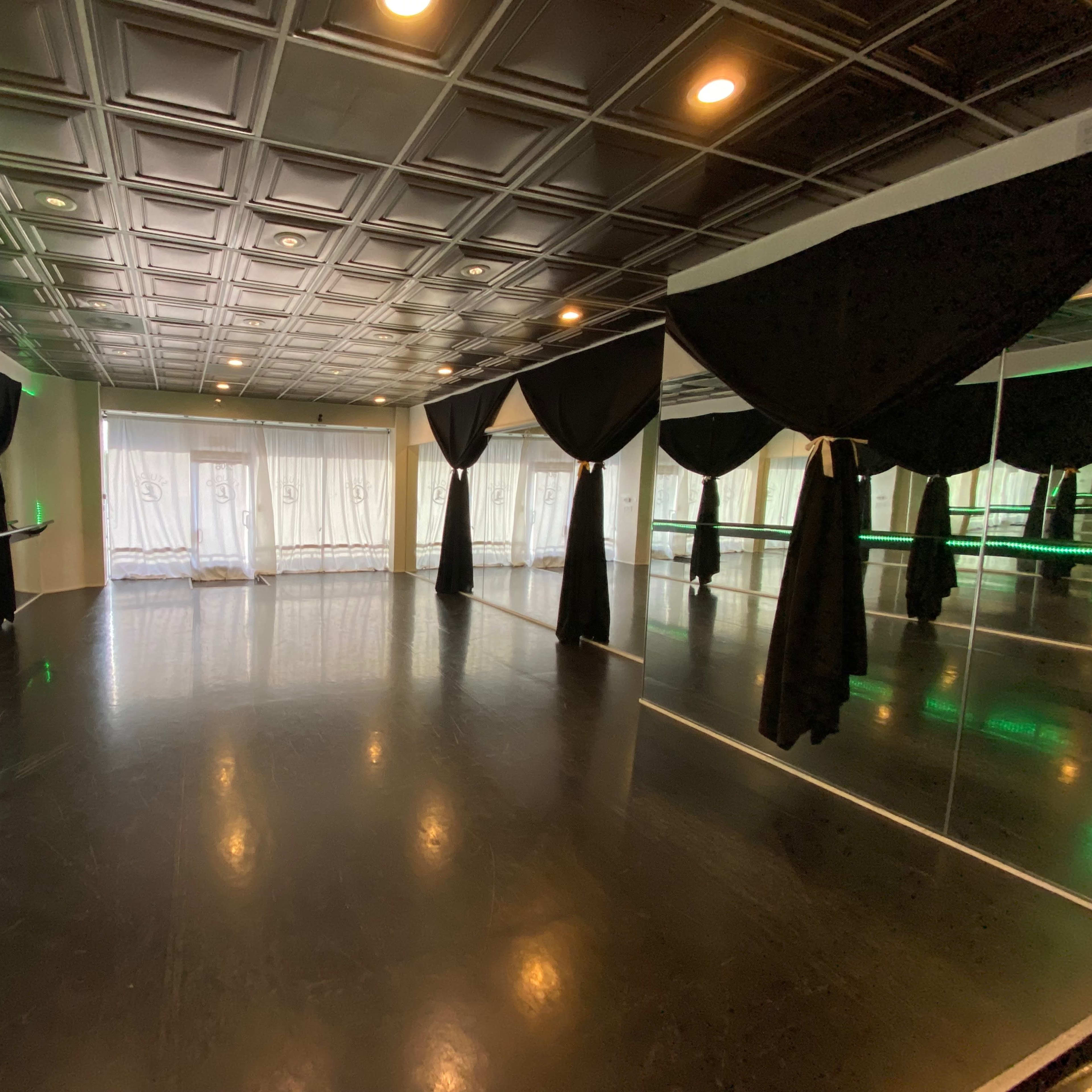 A dance studio with a polished black floor, large windows draped with sheer white curtains, and black curtains hanging from the ceiling.