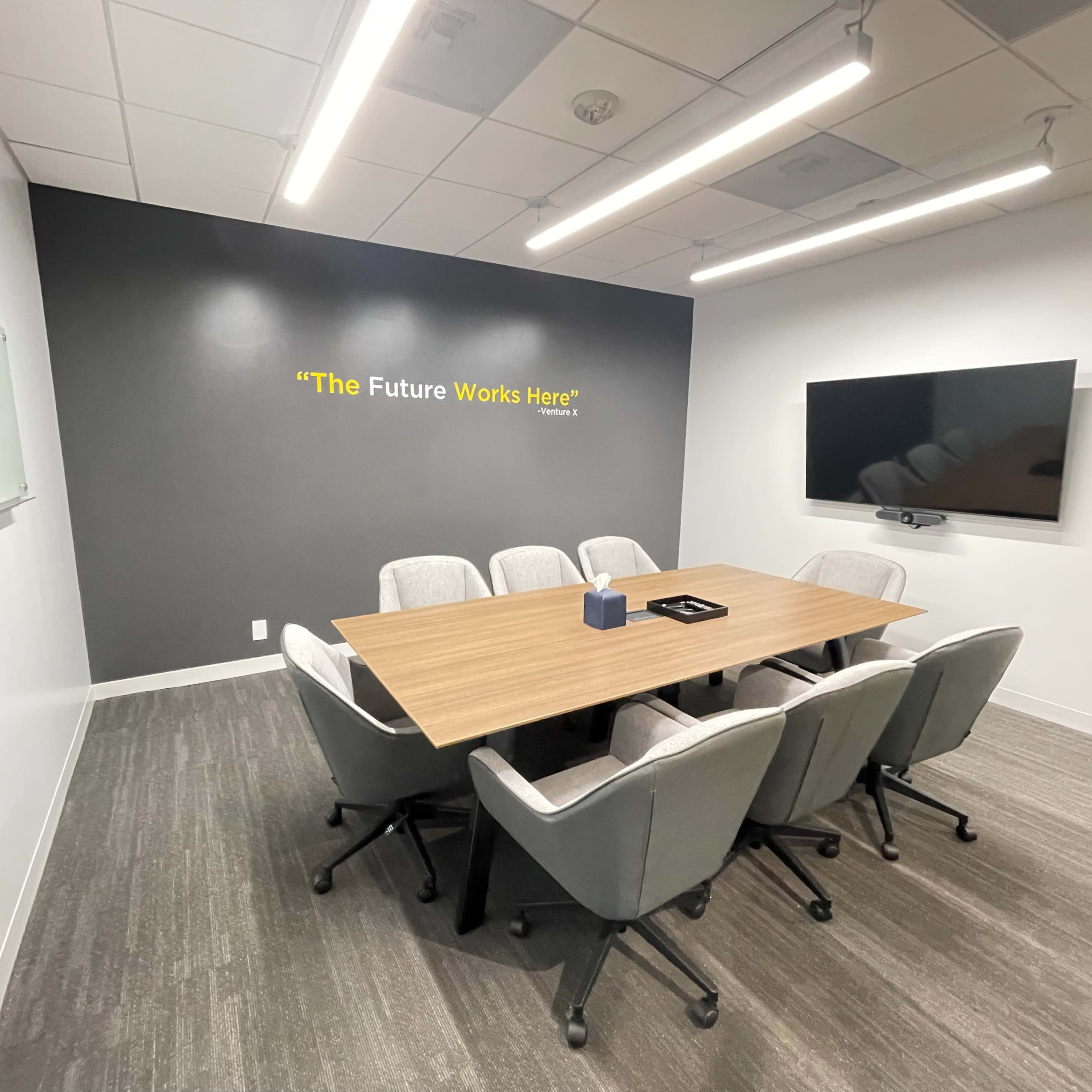 A modern conference room features a large wooden table surrounded by six upholstered chairs, with a wall-mounted screen and a motivational quote displayed above.