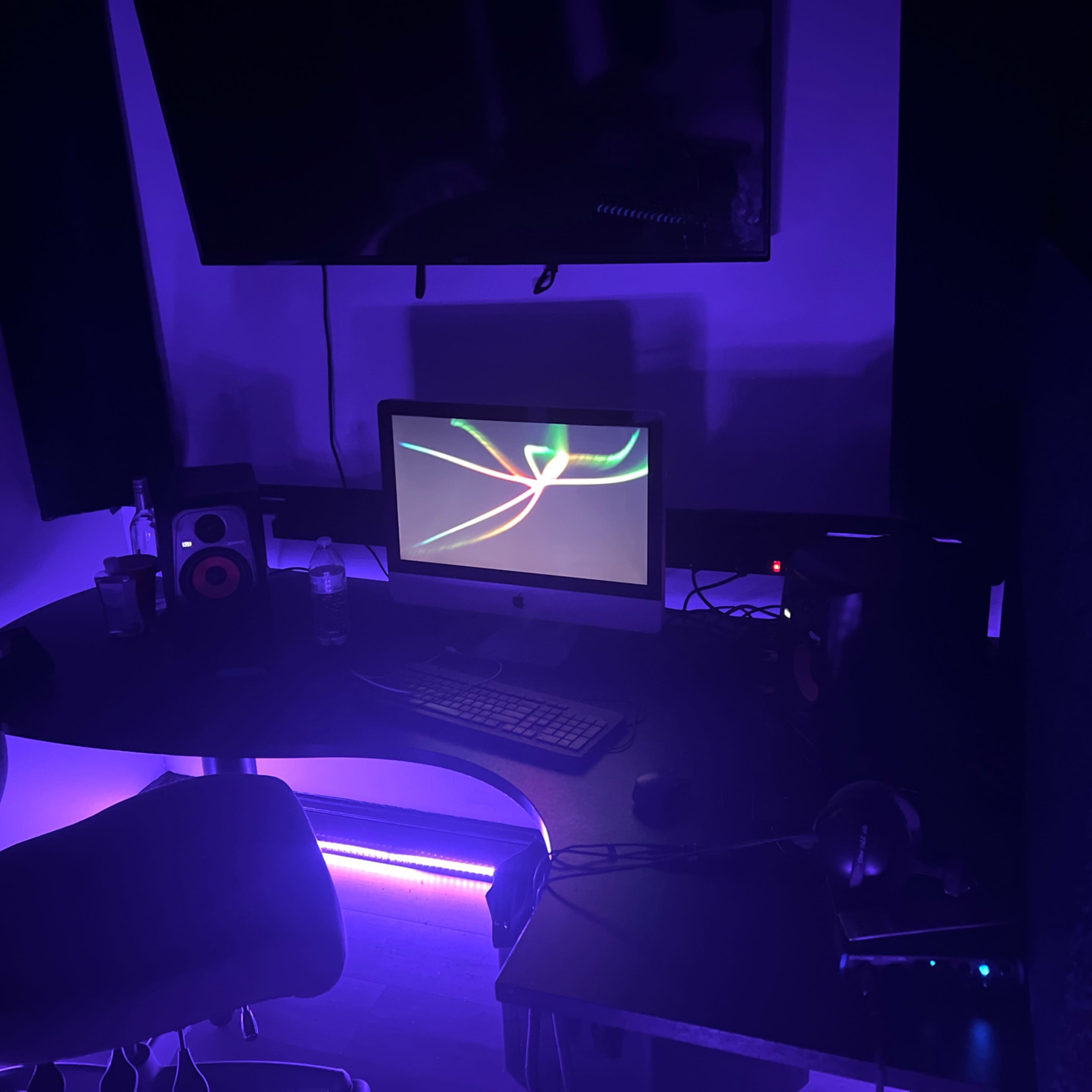 A dimly lit workspace features a computer setup with a monitor, keyboard, speakers, and a chair, surrounded by purple lighting.