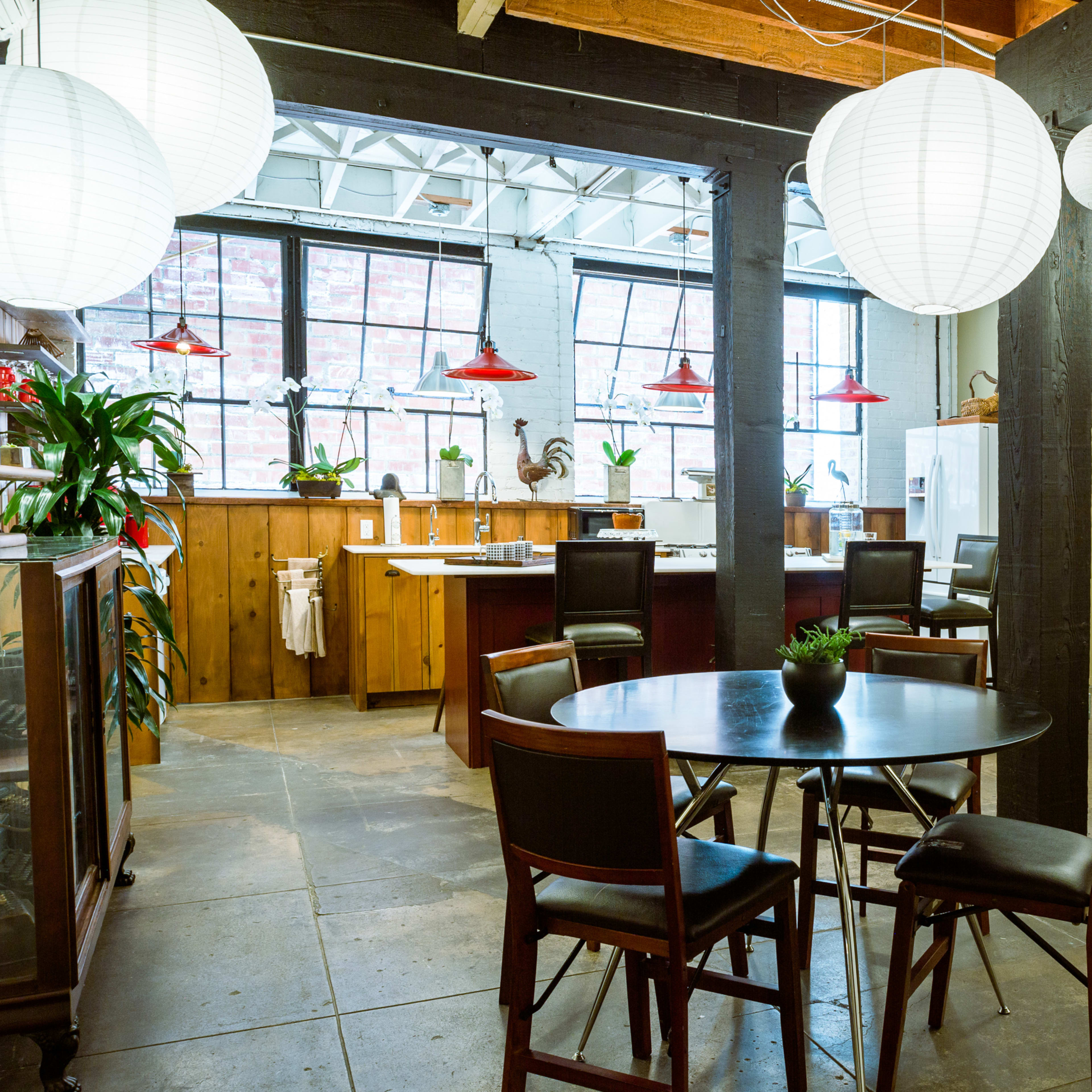A spacious dining area with a round table and chairs, surrounded by a kitchen, pendant lights, and potted plants in a well-lit room.