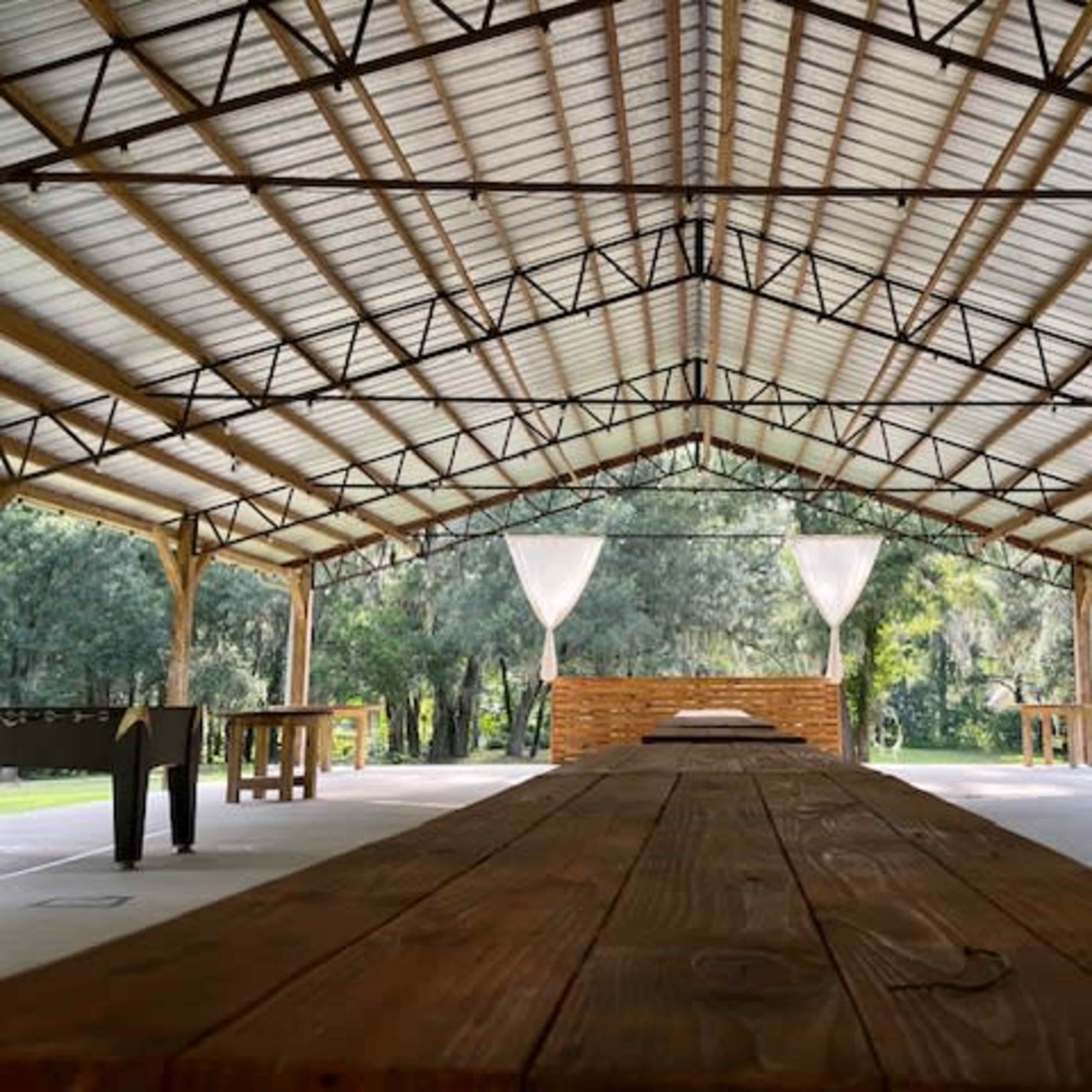 A spacious, open pavilion with wooden beams and a long table set in a green outdoor area.