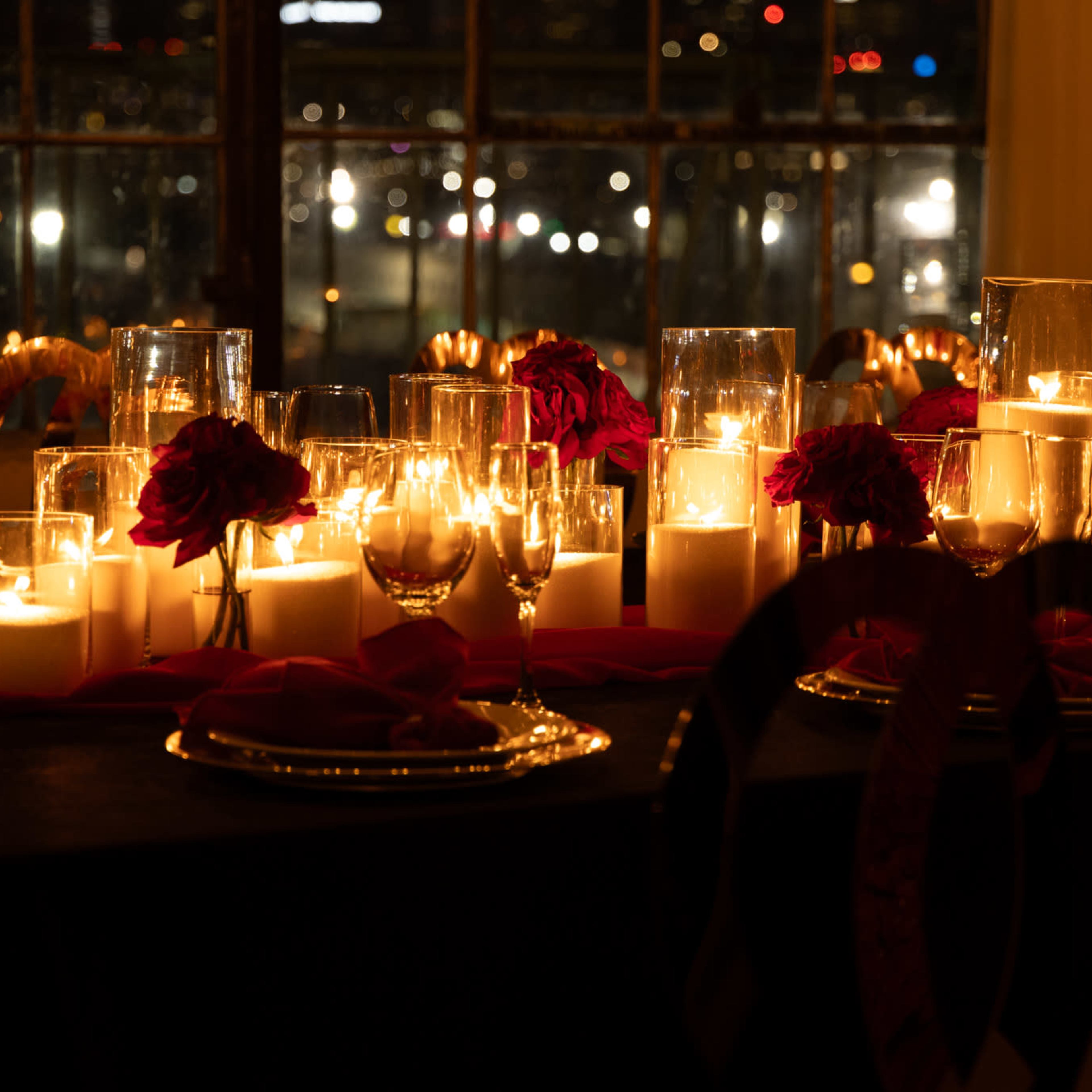 A table is set with numerous candles and red roses, creating a warm glow against a dark backdrop.