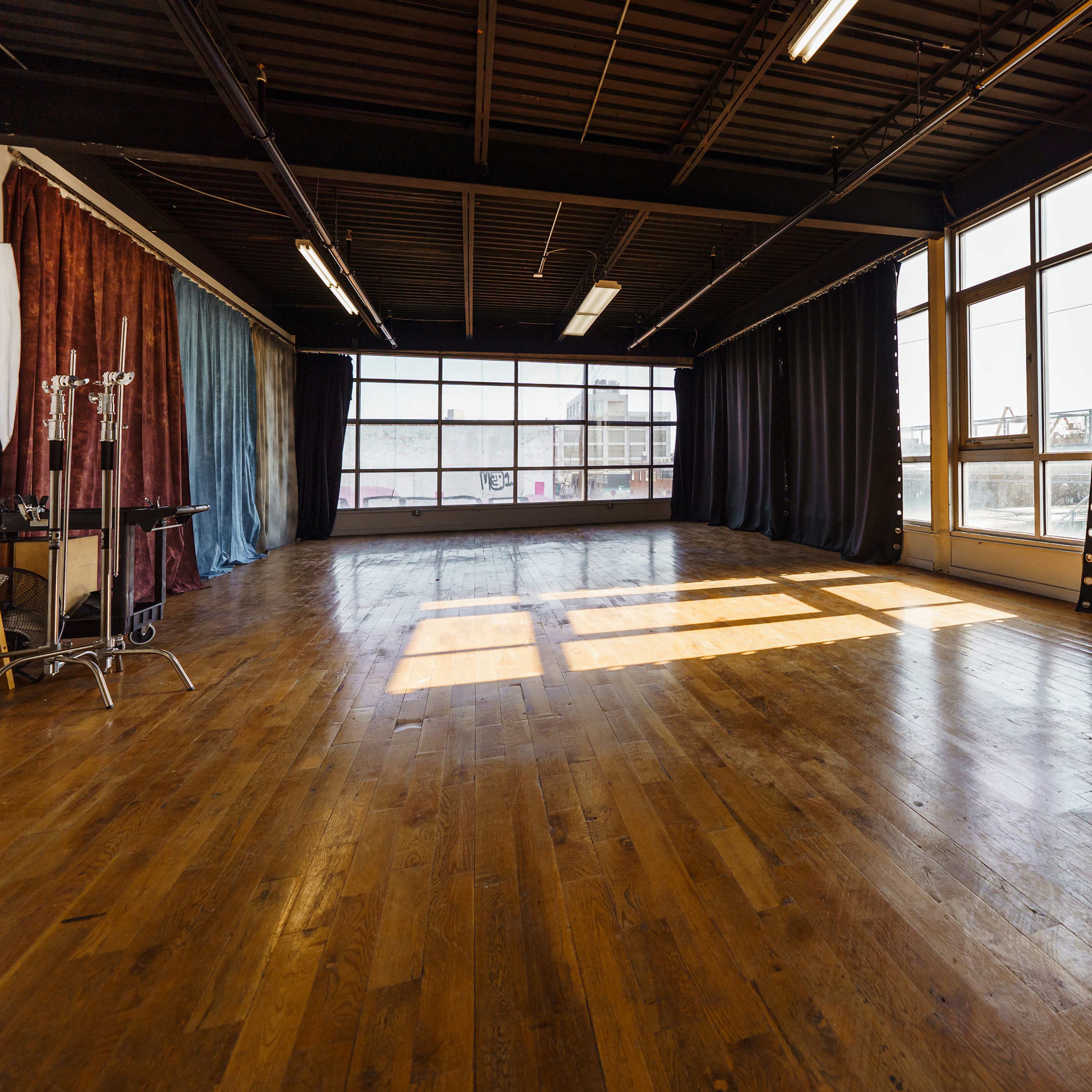 An empty studio space with large windows, wooden floors, and black curtains along the walls.