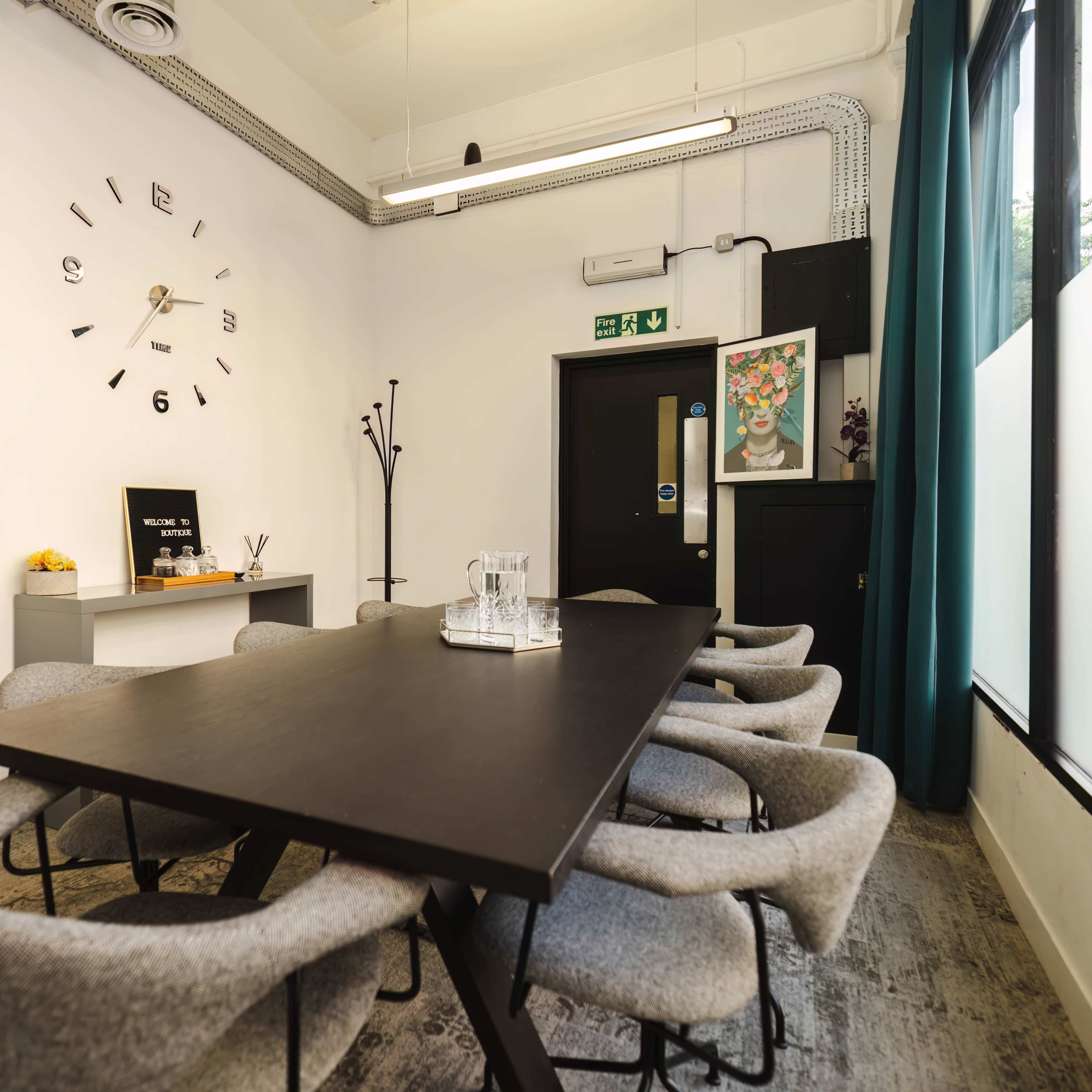 A conference room is equipped with a long black table surrounded by gray chairs, with a wall clock and decorative artwork in the background.