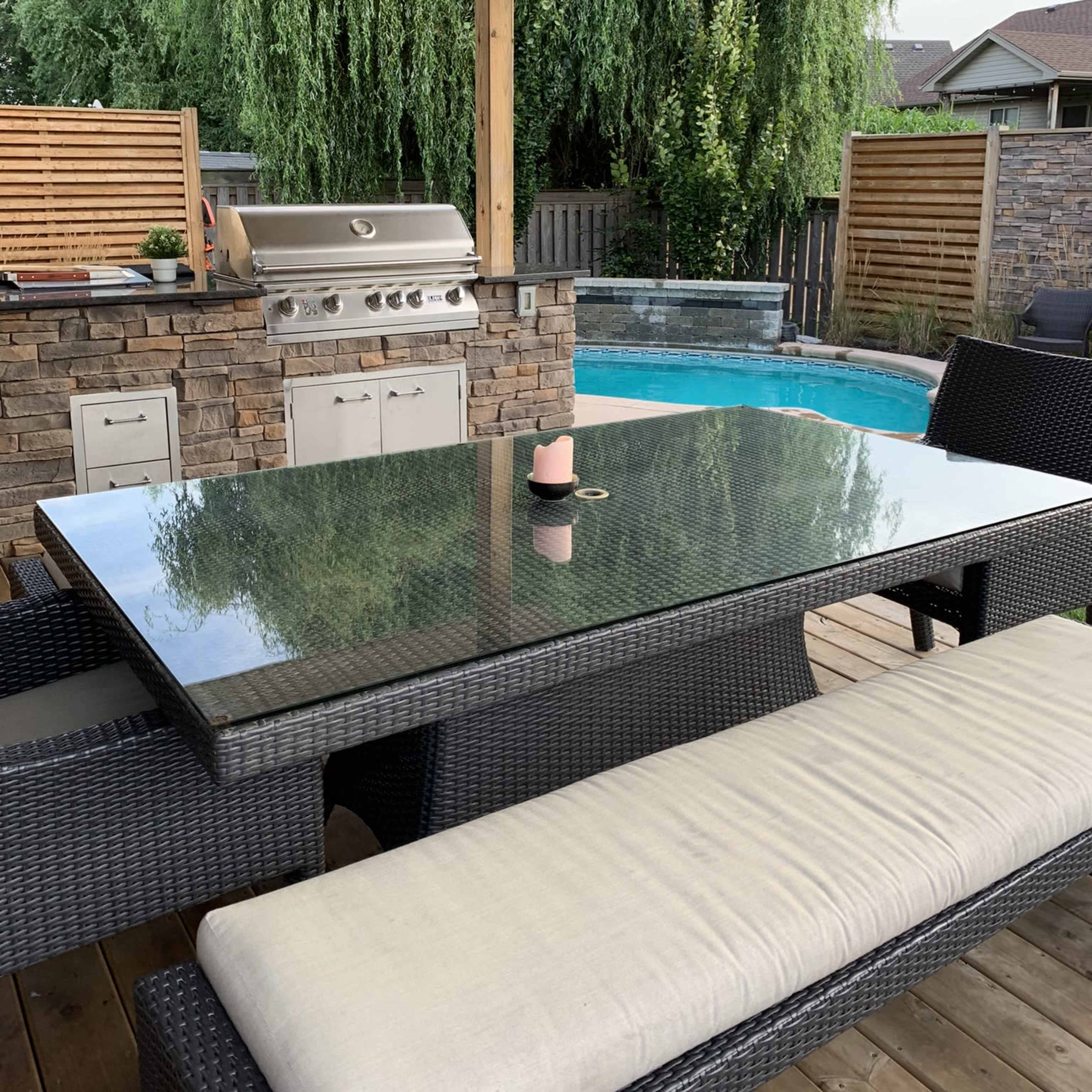 An outdoor patio features a stone barbecue grill, a glass-top dining table with seating, and a pool visible in the background.