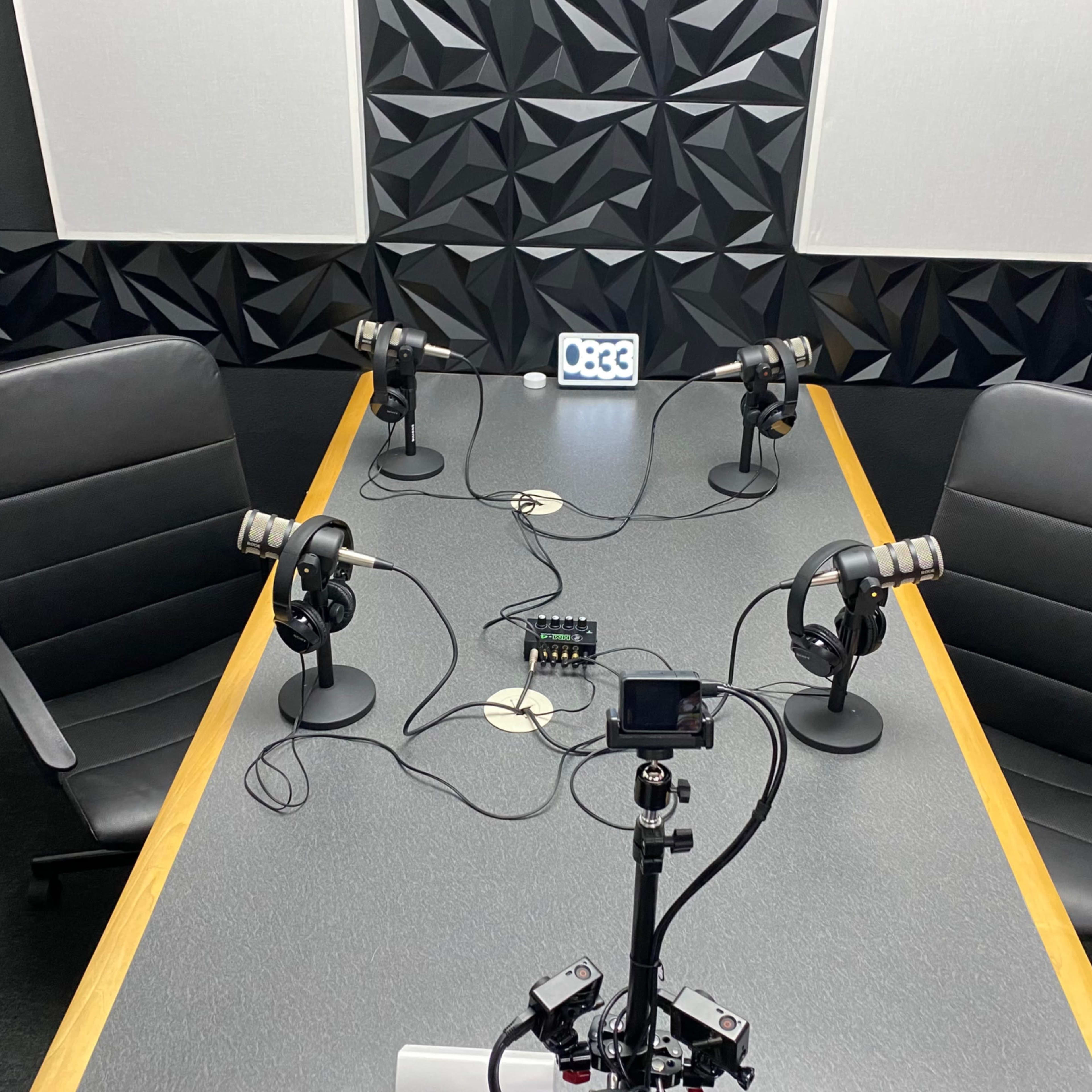 A podcast recording studio features a rectangular table with four microphones, two chairs, and a device for audio mixing, set against a textured black wall.