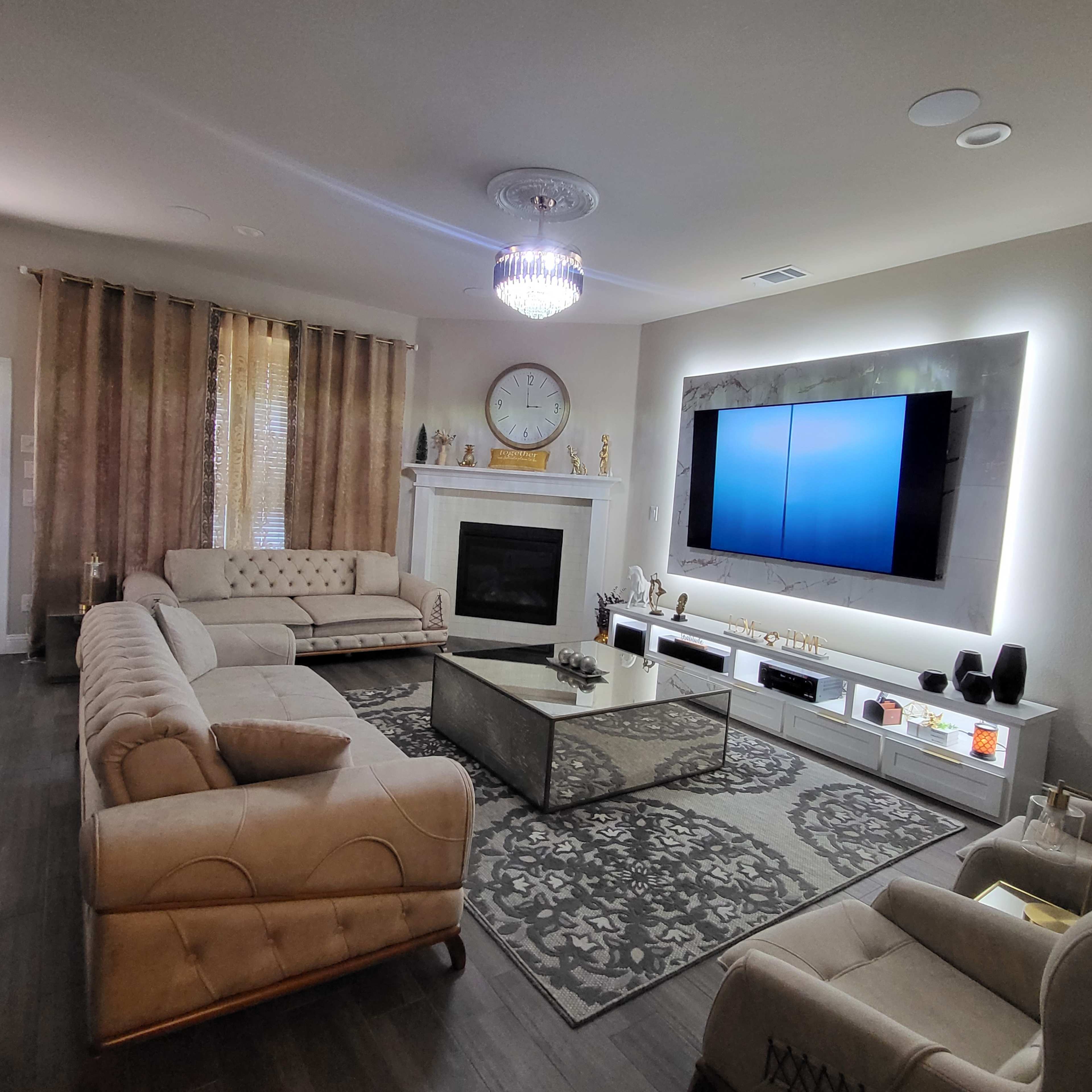 The living room features a modern design with a large flat-screen TV mounted above a sleek entertainment unit, a central coffee table, and beige seating arranged around a decorative area rug.