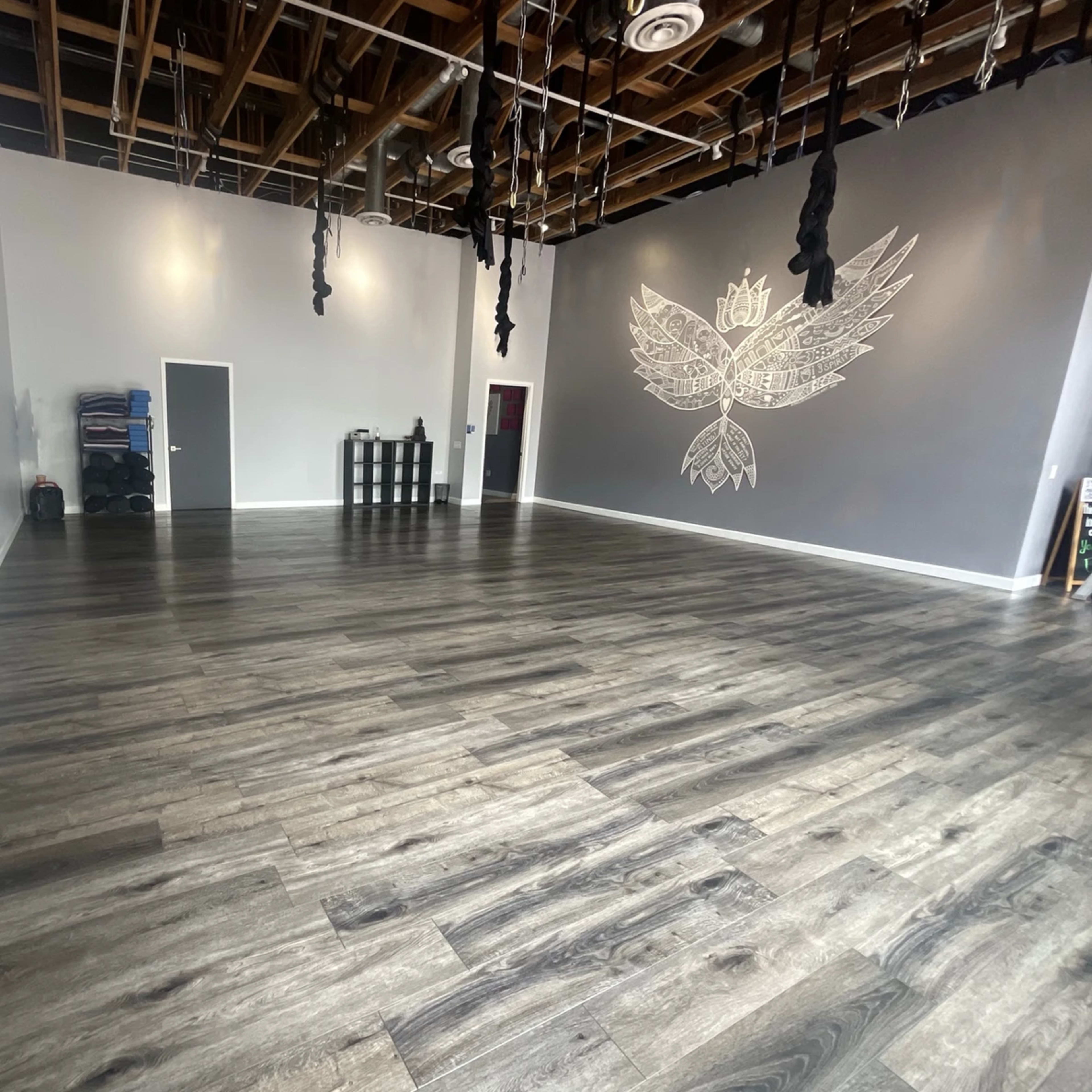 A spacious, empty studio with wooden flooring, white walls, and a gray accent wall featuring a large design, along with some equipment in the corner.