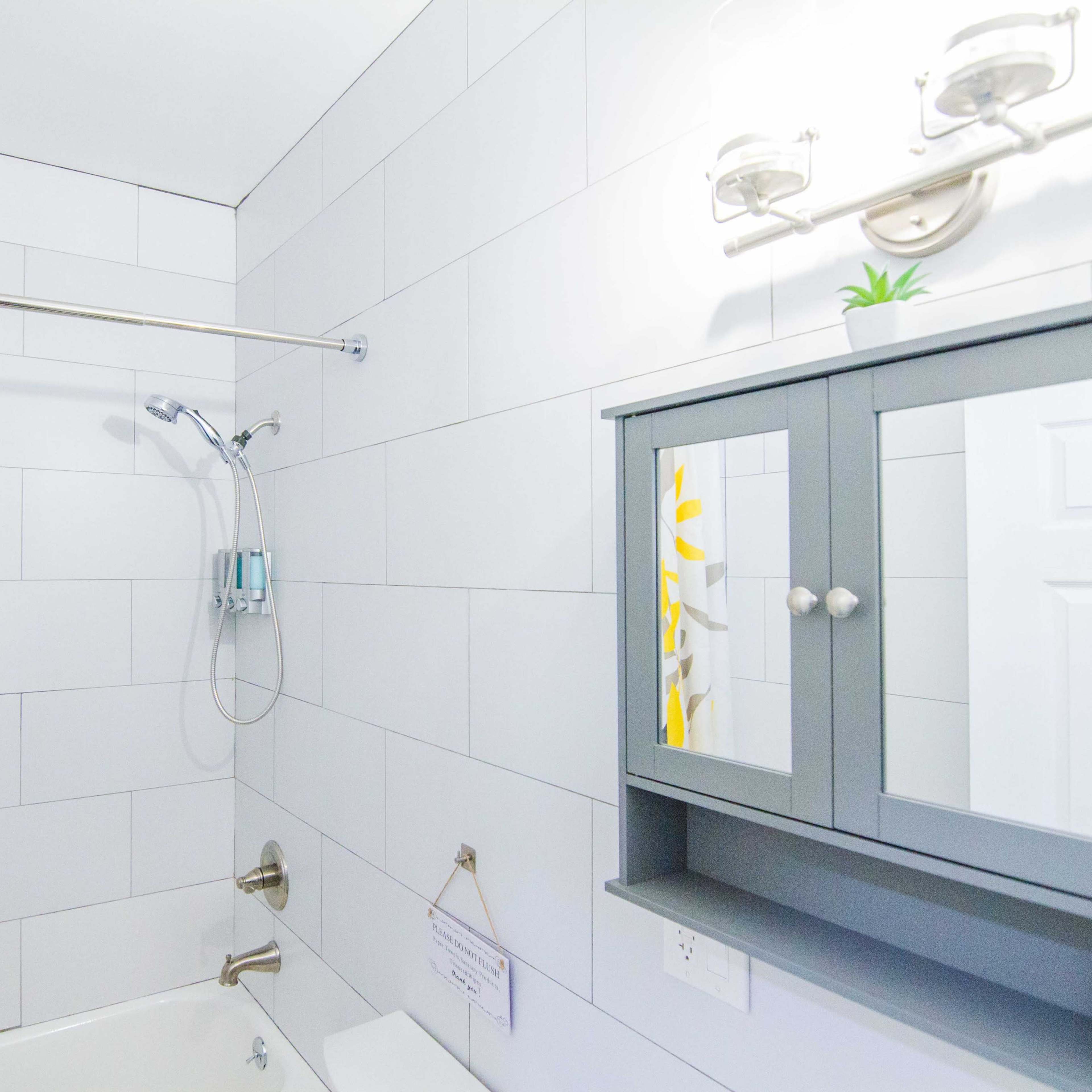 A bathroom featuring a bathtub with a shower, light gray tiled walls, and a mirror cabinet above a sink, complemented by a shower curtain with a yellow floral design.