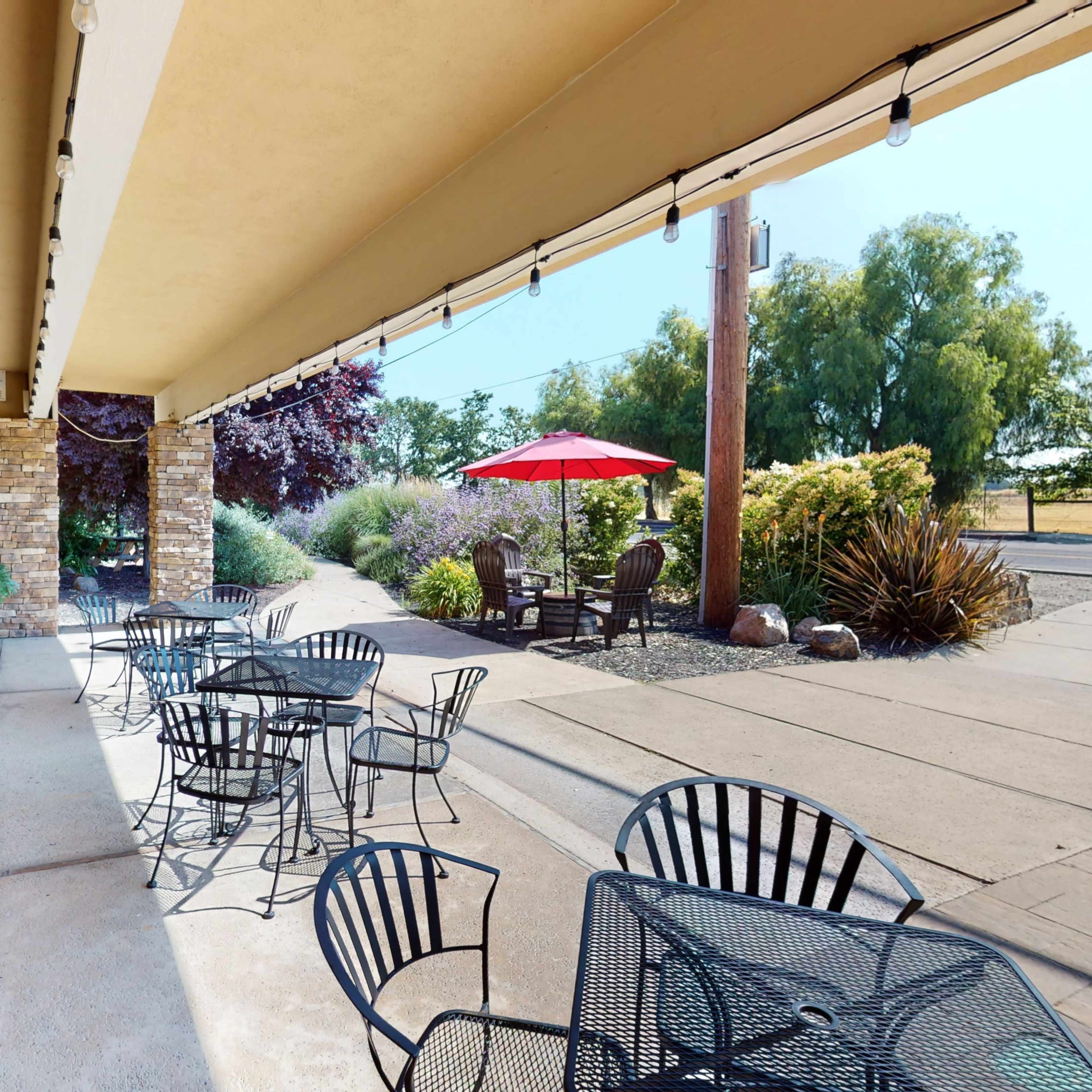 A café exterior with outdoor seating, featuring black metal tables and chairs, along with a chalkboard sign and an umbrella over one table, set against a backdrop of landscaped greenery.