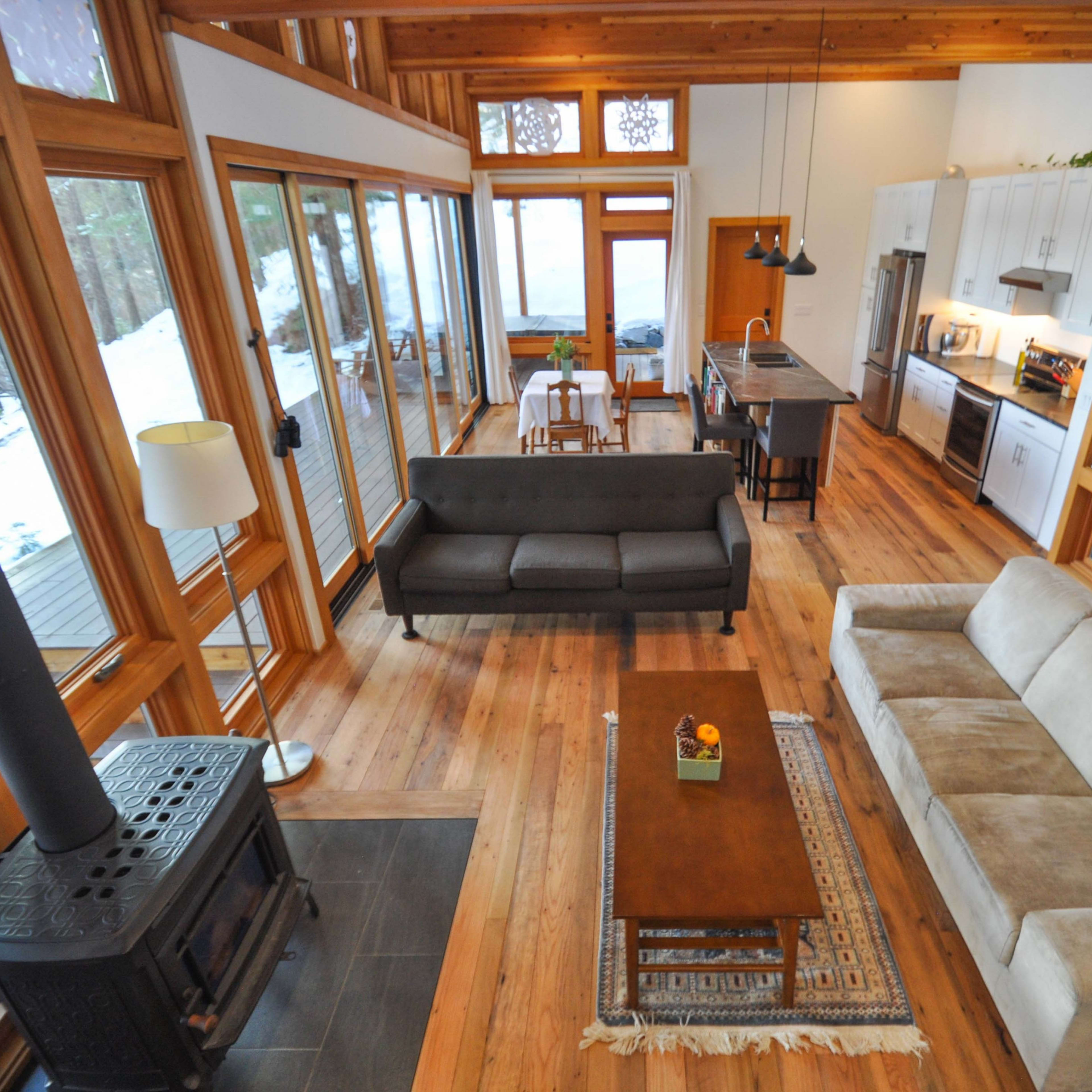 A spacious, modern living room features large windows, wooden beams, and a mix of seating arrangements with a kitchen area visible in the background.