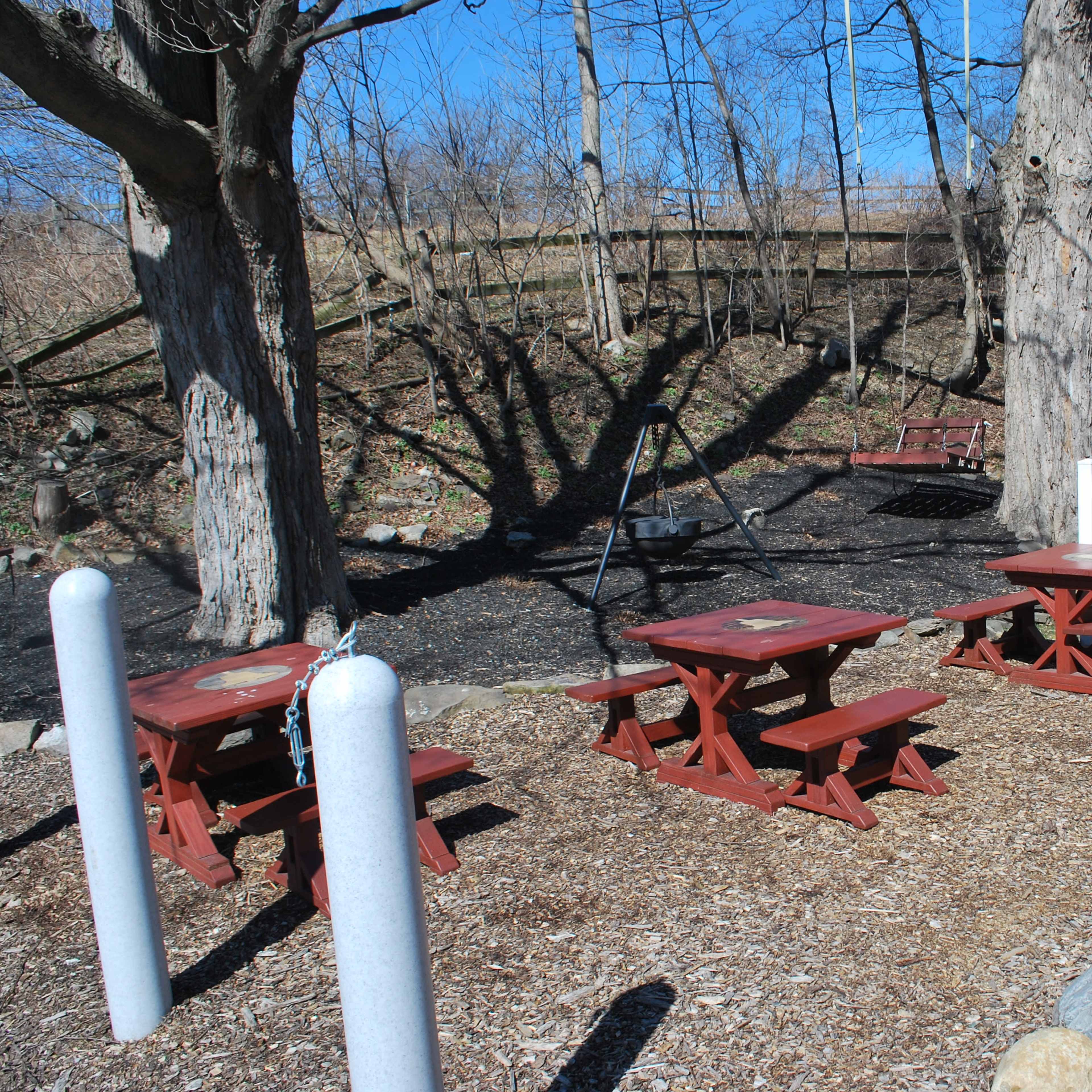 A rustic outdoor seating area with wooden picnic tables surrounded by trees and a white picket fence.