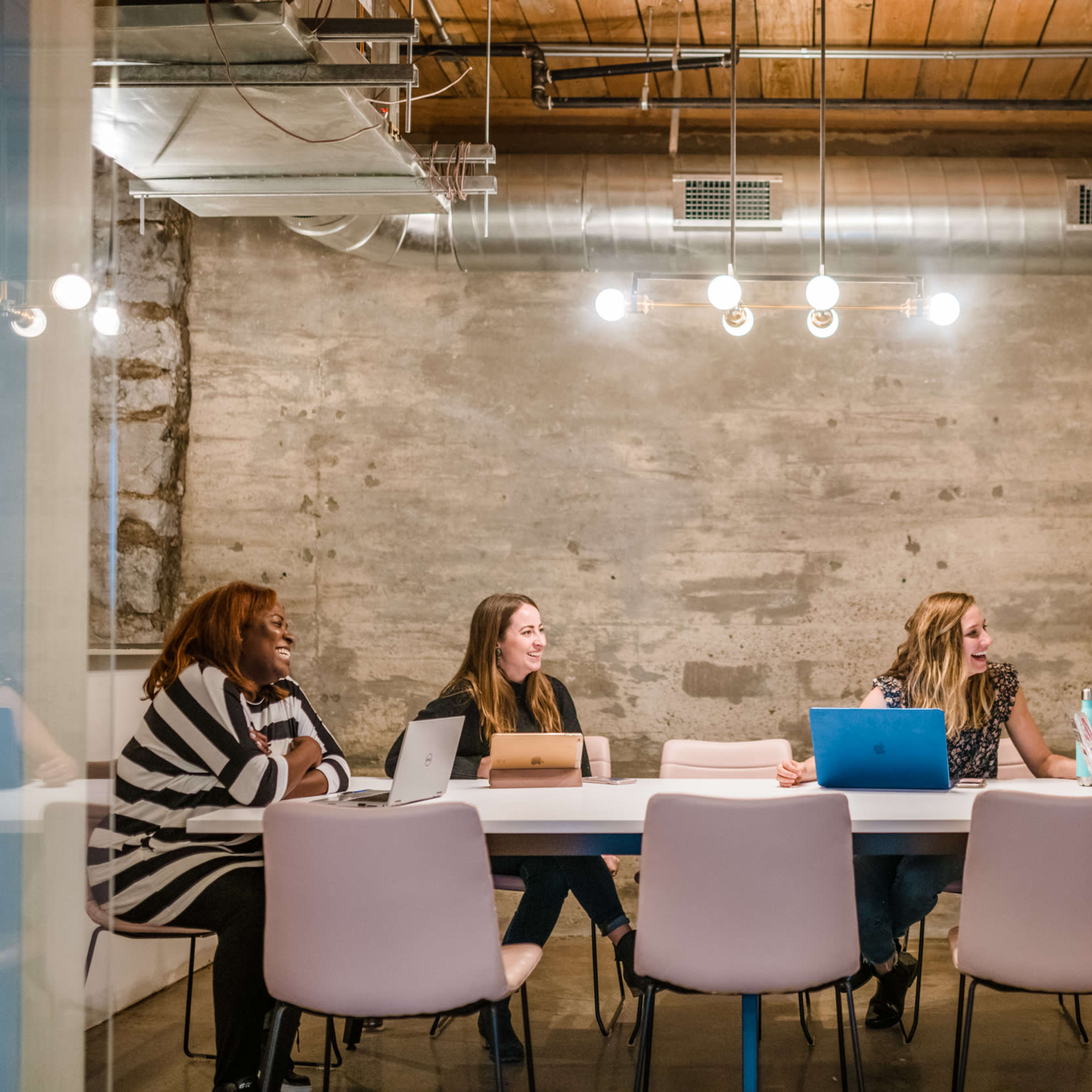 A group of five women is seated around a table in a modern meeting room with a concrete wall and overhead lighting.