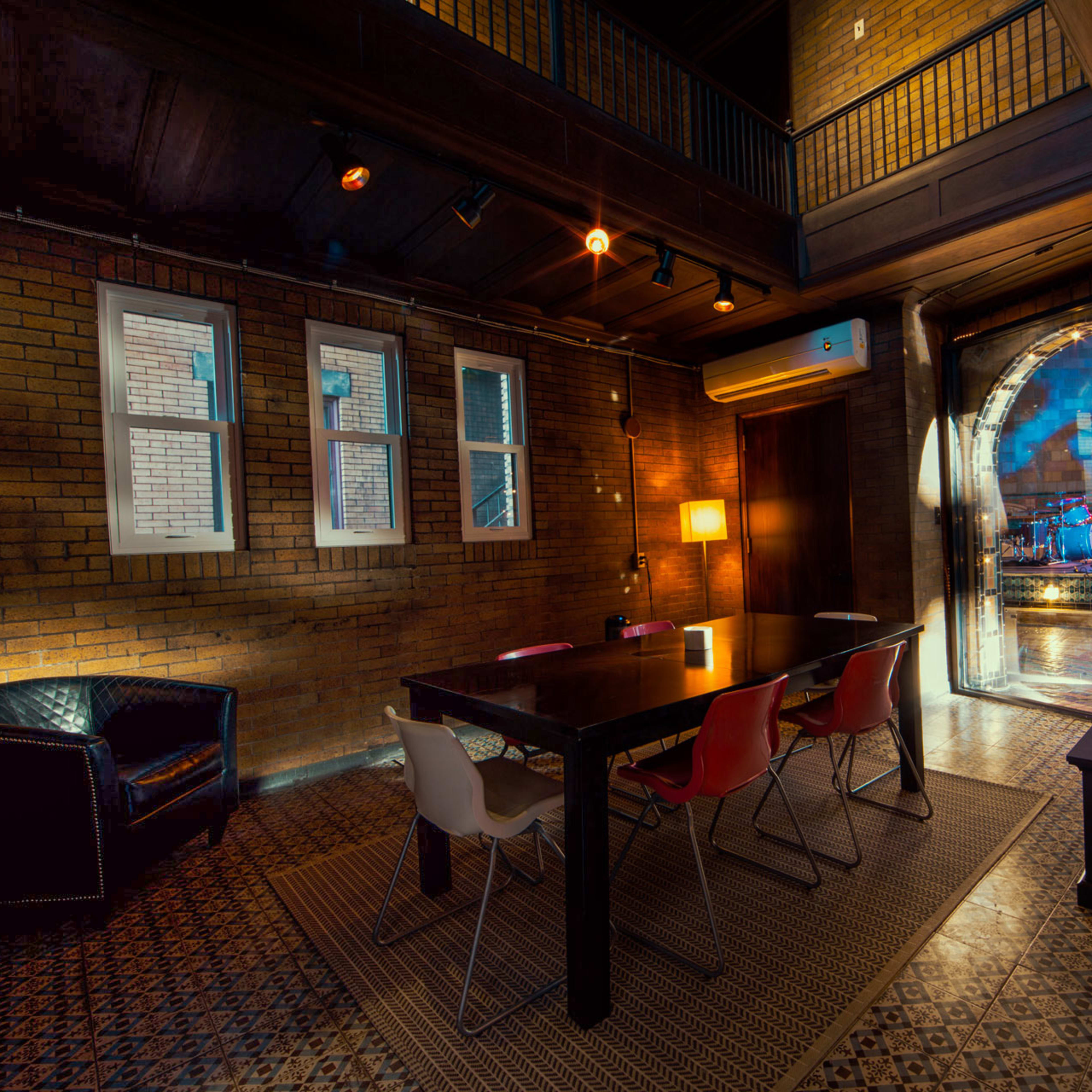 A dimly lit room with exposed brick walls, featuring a large wooden table surrounded by colorful chairs, and an archway leading to another space.