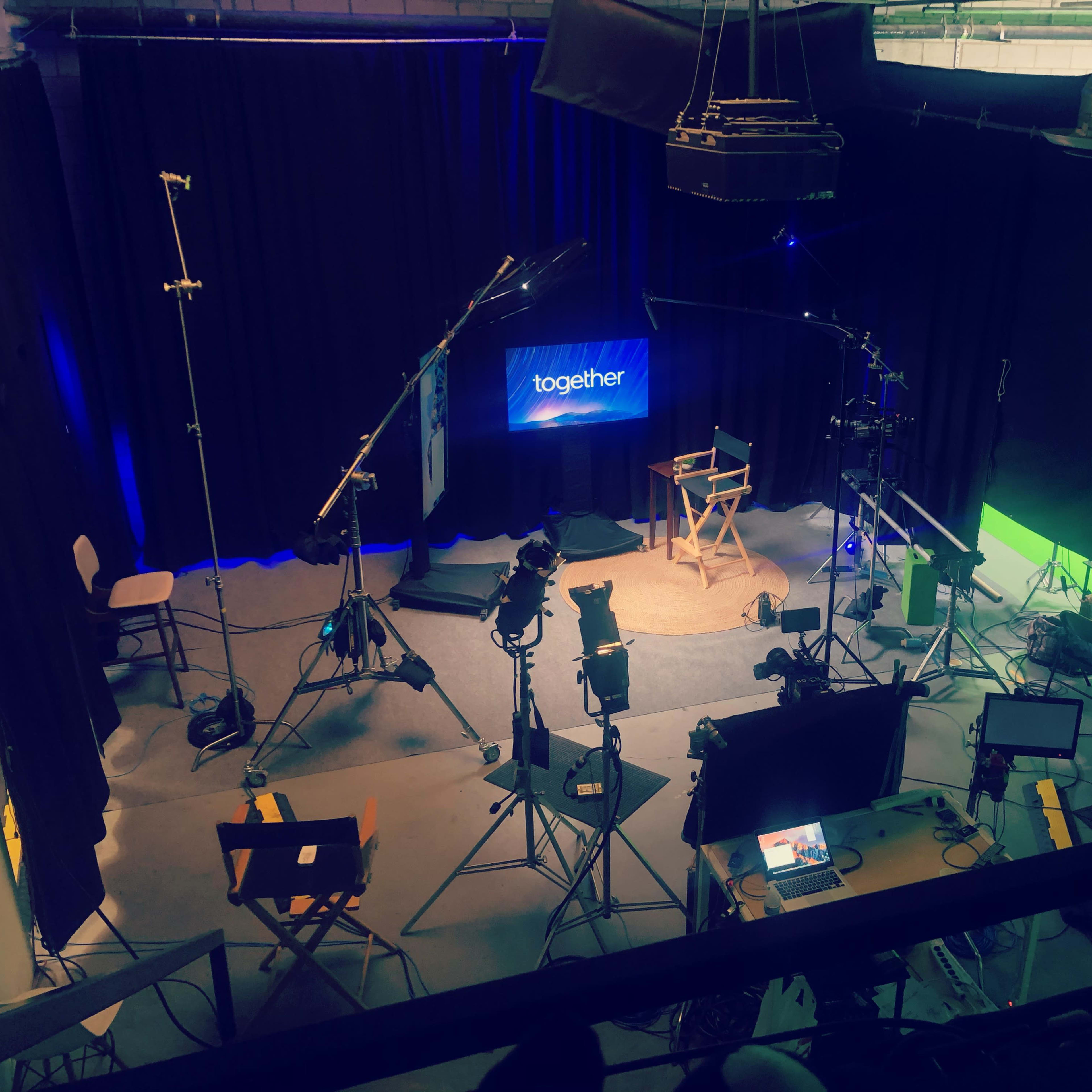 A film studio setup features a central director's chair, lighting equipment, cameras, and a green screen backdrop.