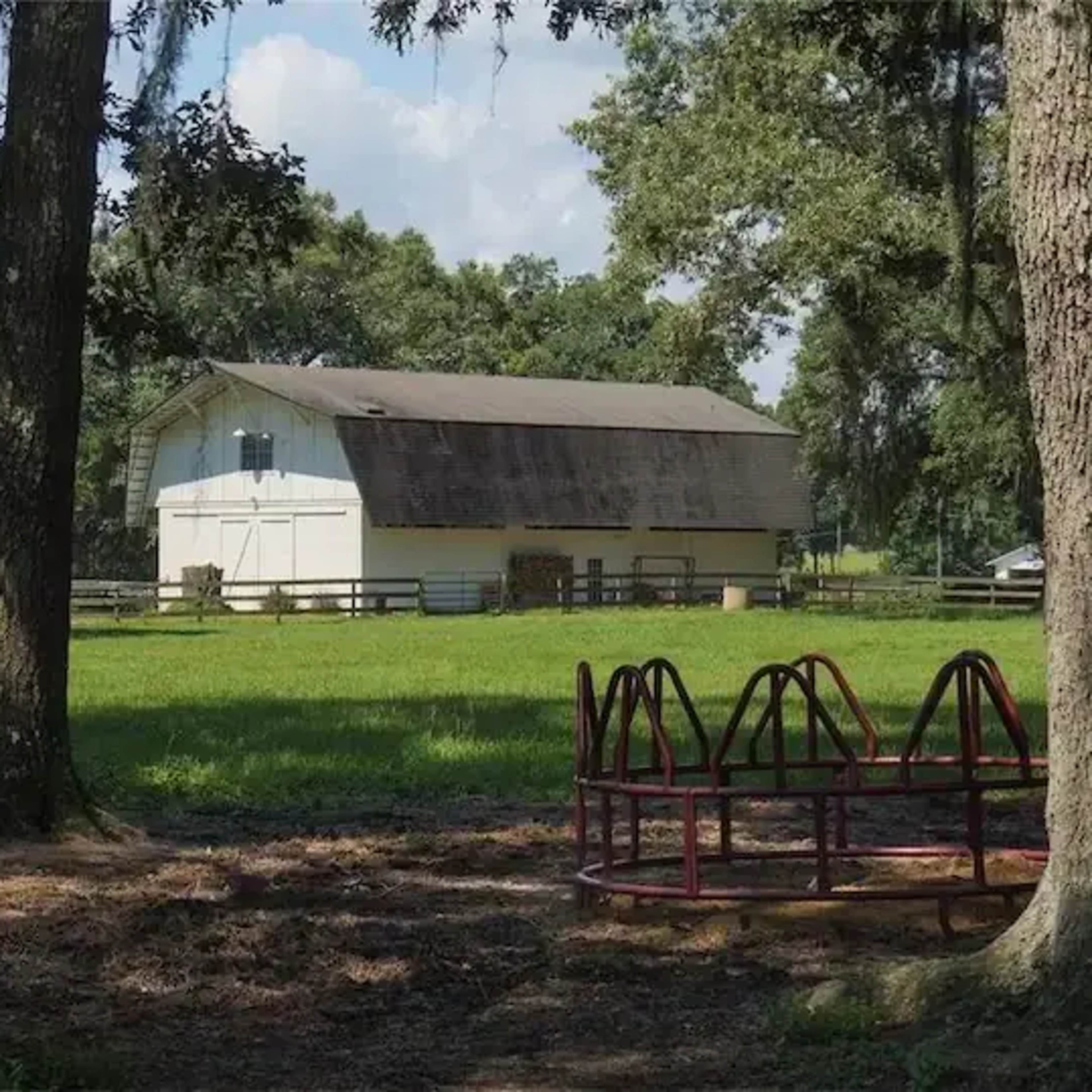 A white barn stands in a grassy field, surrounded by trees, with a playground structure in the foreground.