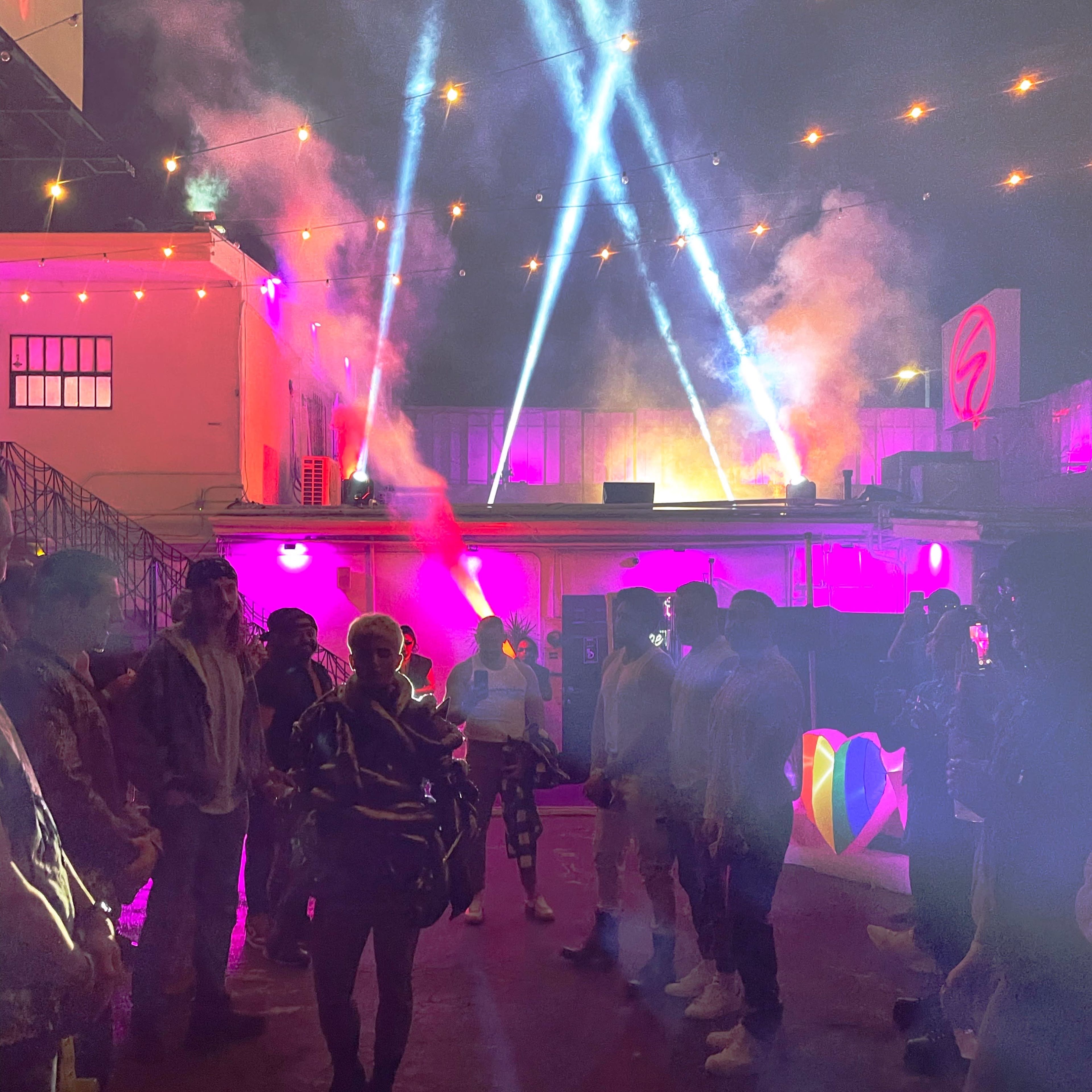 A crowd of people stands in a lit outdoor space with colorful lights and fog effects, while beams of light shine down from above.