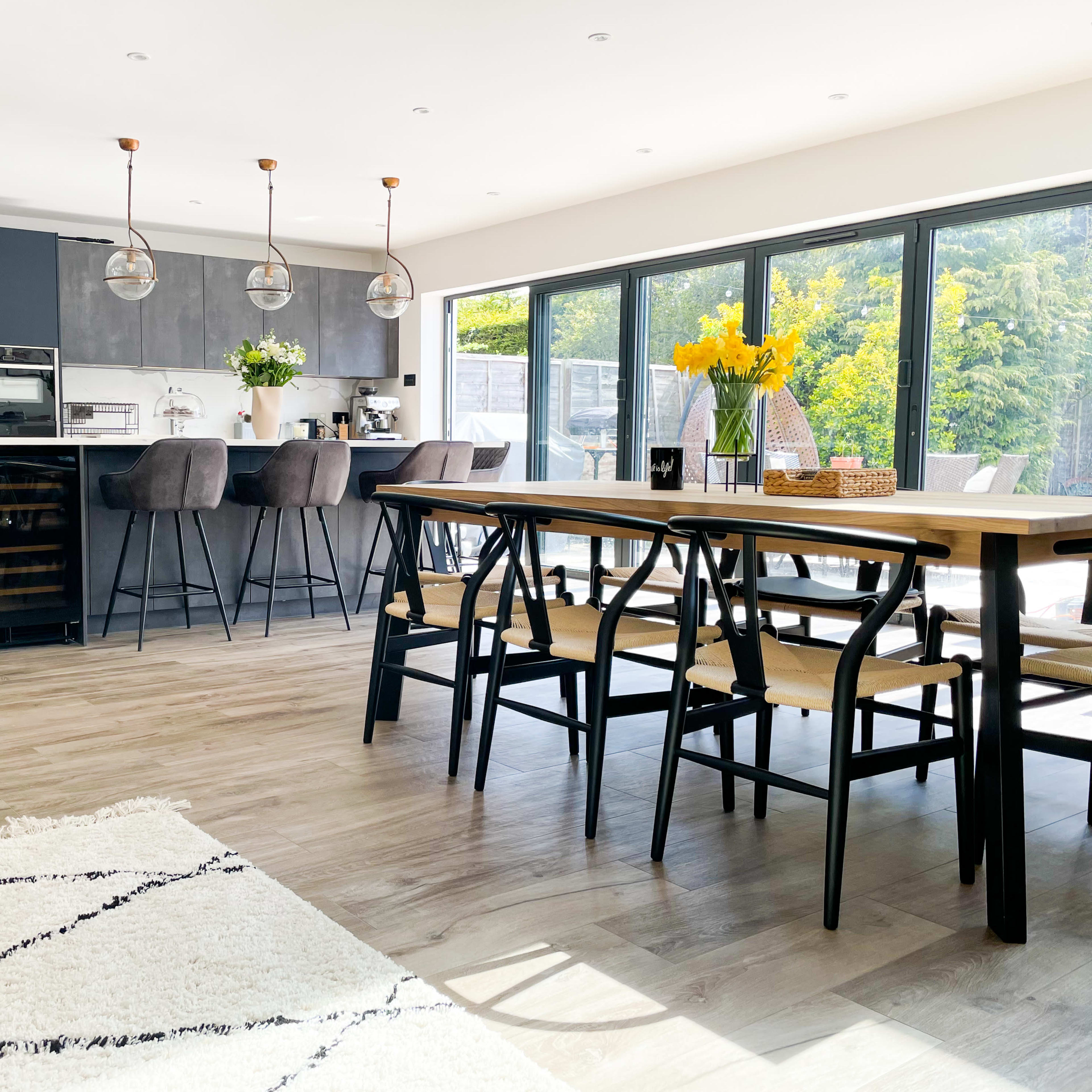 A modern kitchen and dining area features a large wooden table, dark cabinetry, and large windows overlooking a garden.