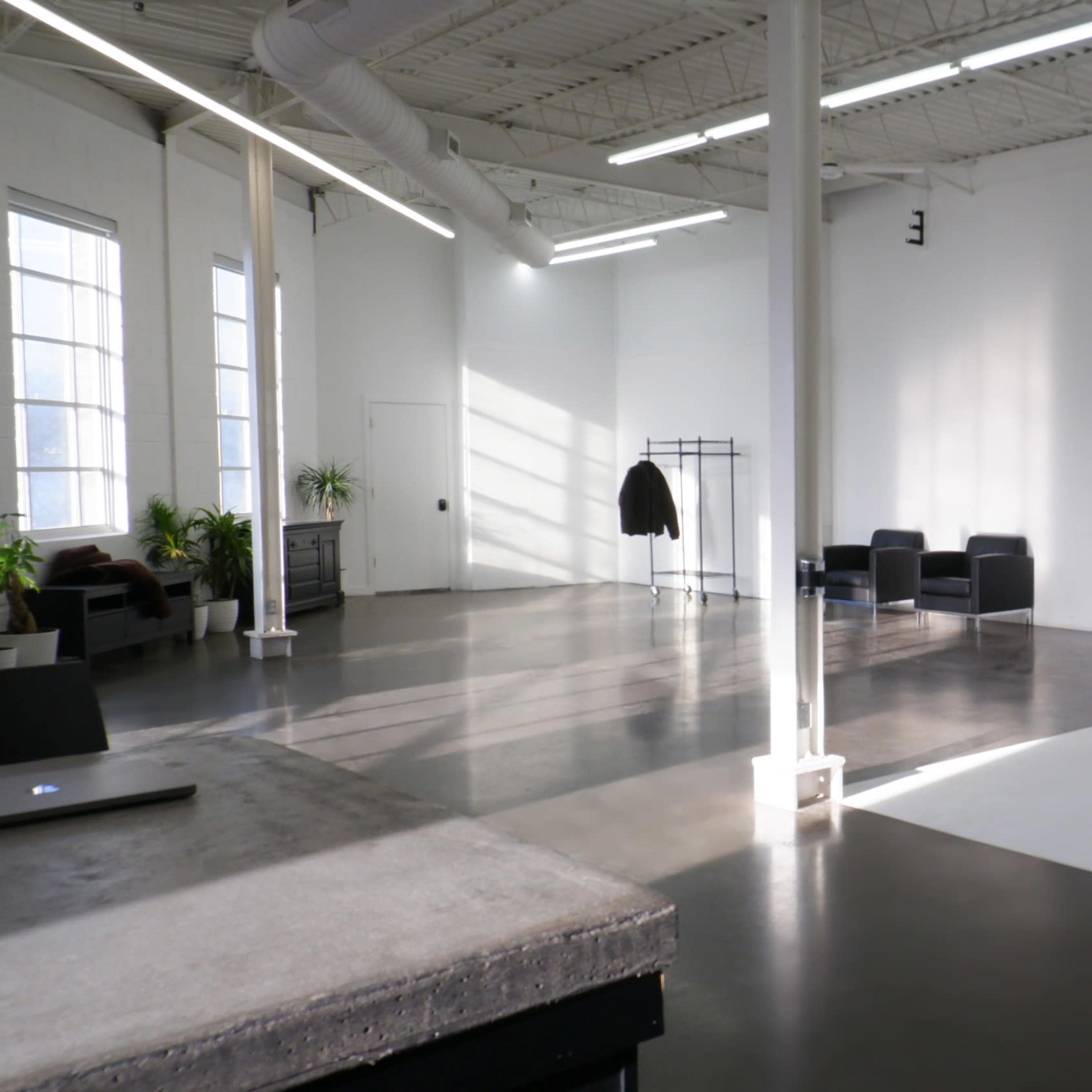 A spacious, bright studio with a polished concrete floor, a white backdrop, and minimalist furniture including a coat rack and two black chairs.