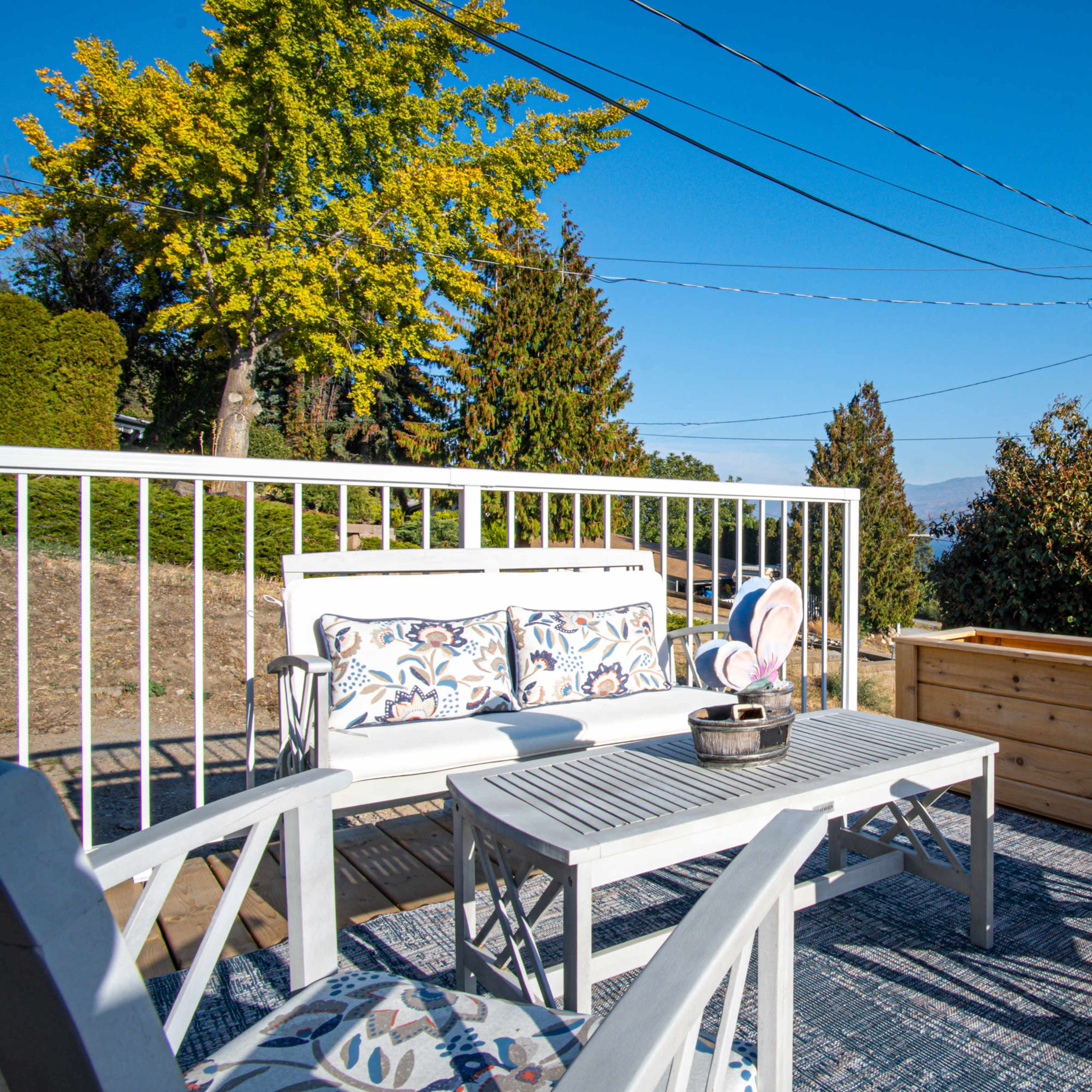 A sunny outdoor seating area features white furniture and a wooden planter, surrounded by trees and mountains under a clear blue sky.