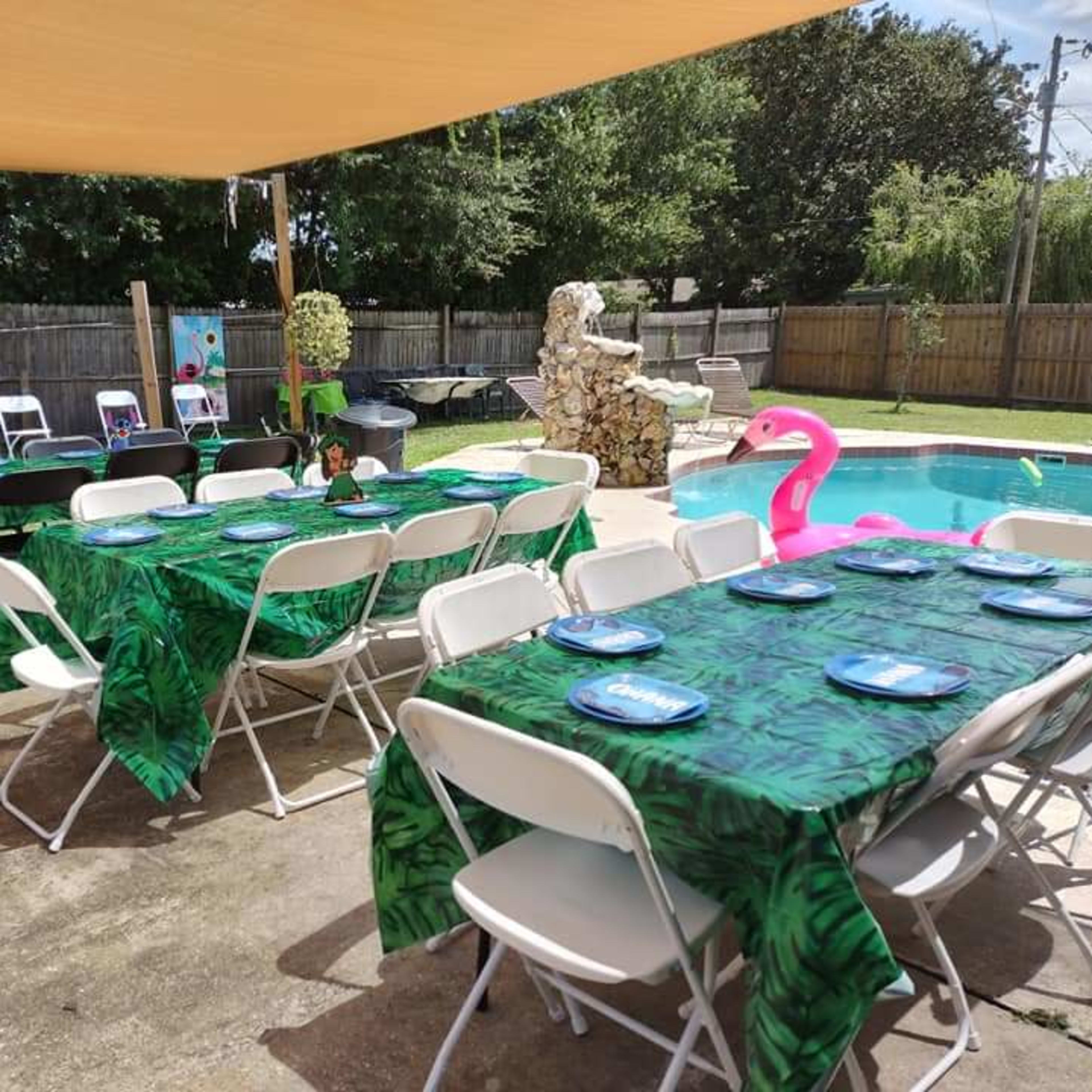 A backyard setup with multiple tables covered in leaf-patterned tablecloths, arranged around a pool area adorned with a pink flamingo and festive decorations.