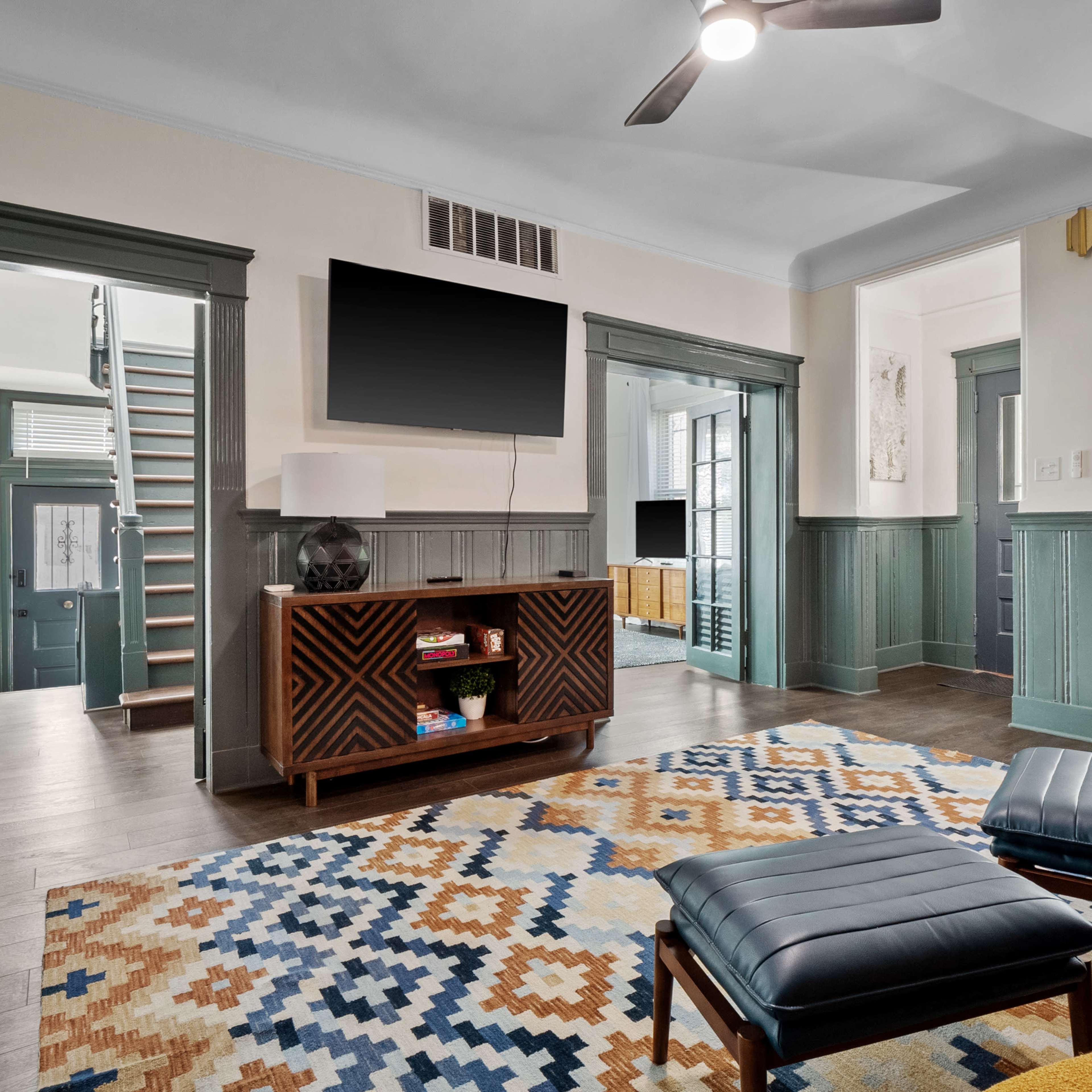 The room features a patterned rug, modern furniture, a mounted television, and a view into an adjacent room with a staircase.