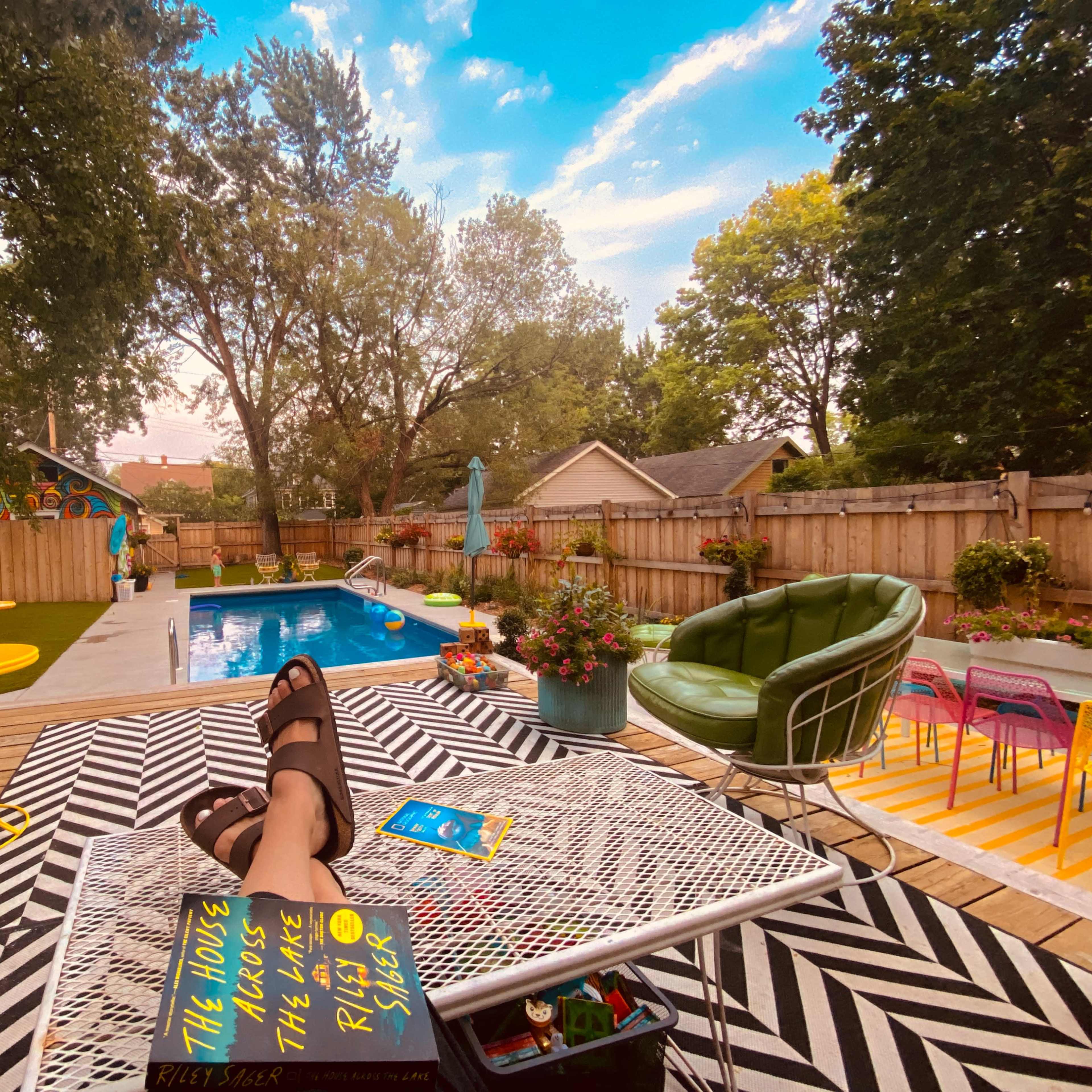 A backyard scene features a swimming pool surrounded by colorful lounge furniture and potted plants, with a person standing nearby and a small dog near the camera.