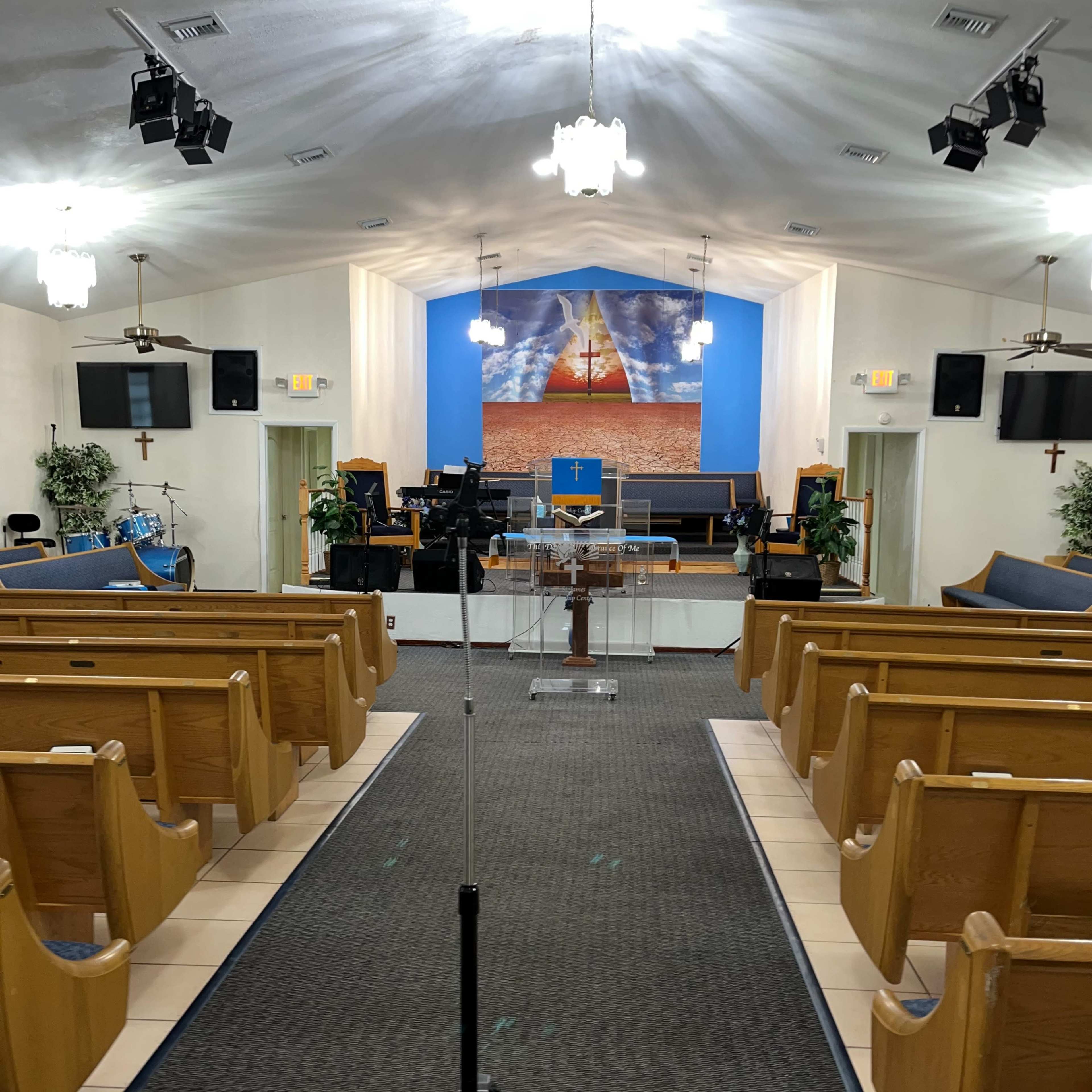 The interior of a church, featuring wooden pews facing a stage with a blue backdrop, a large cross, and musical equipment.