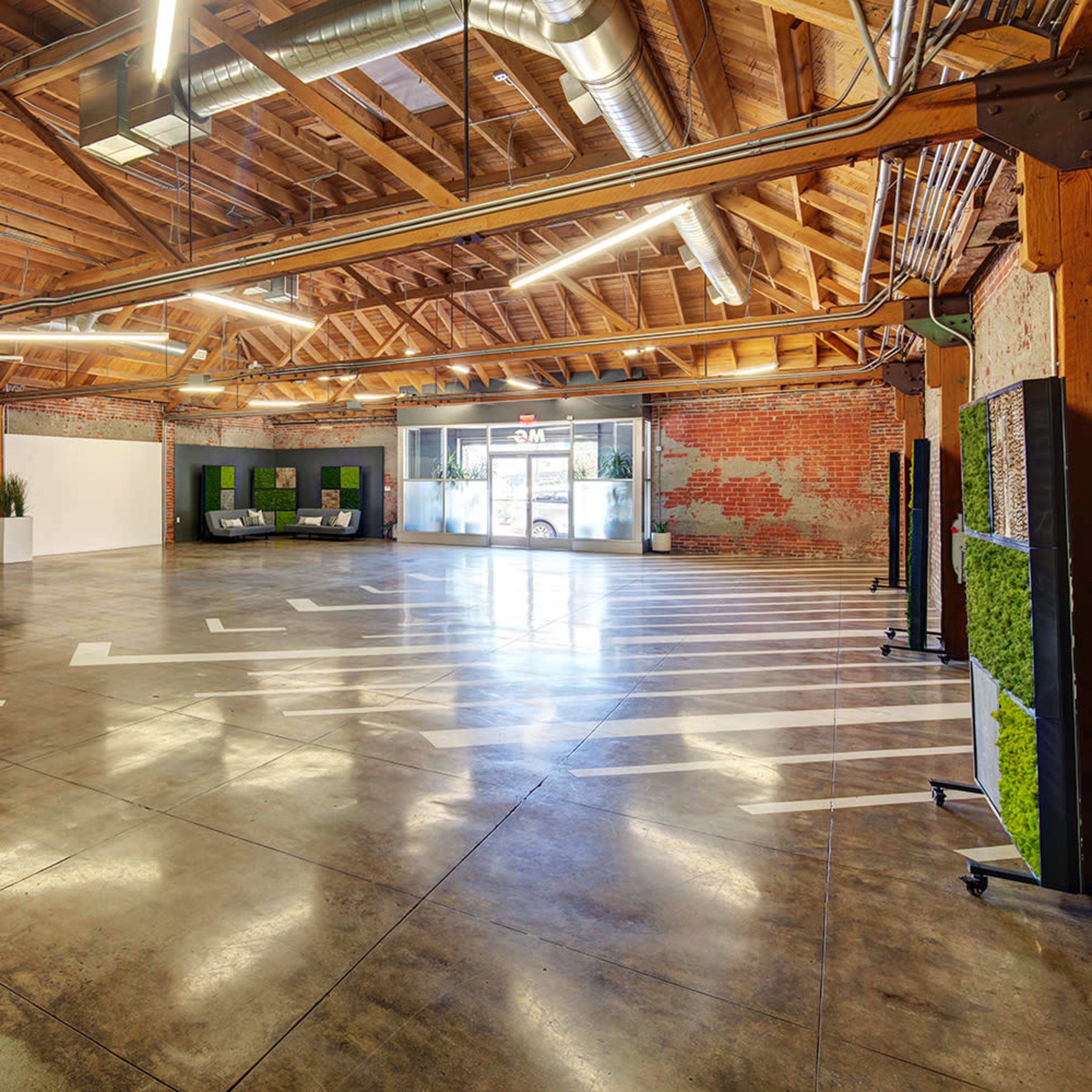 A spacious indoor area with a polished concrete floor, wooden beams, and large windows, featuring minimal furniture and greenery.