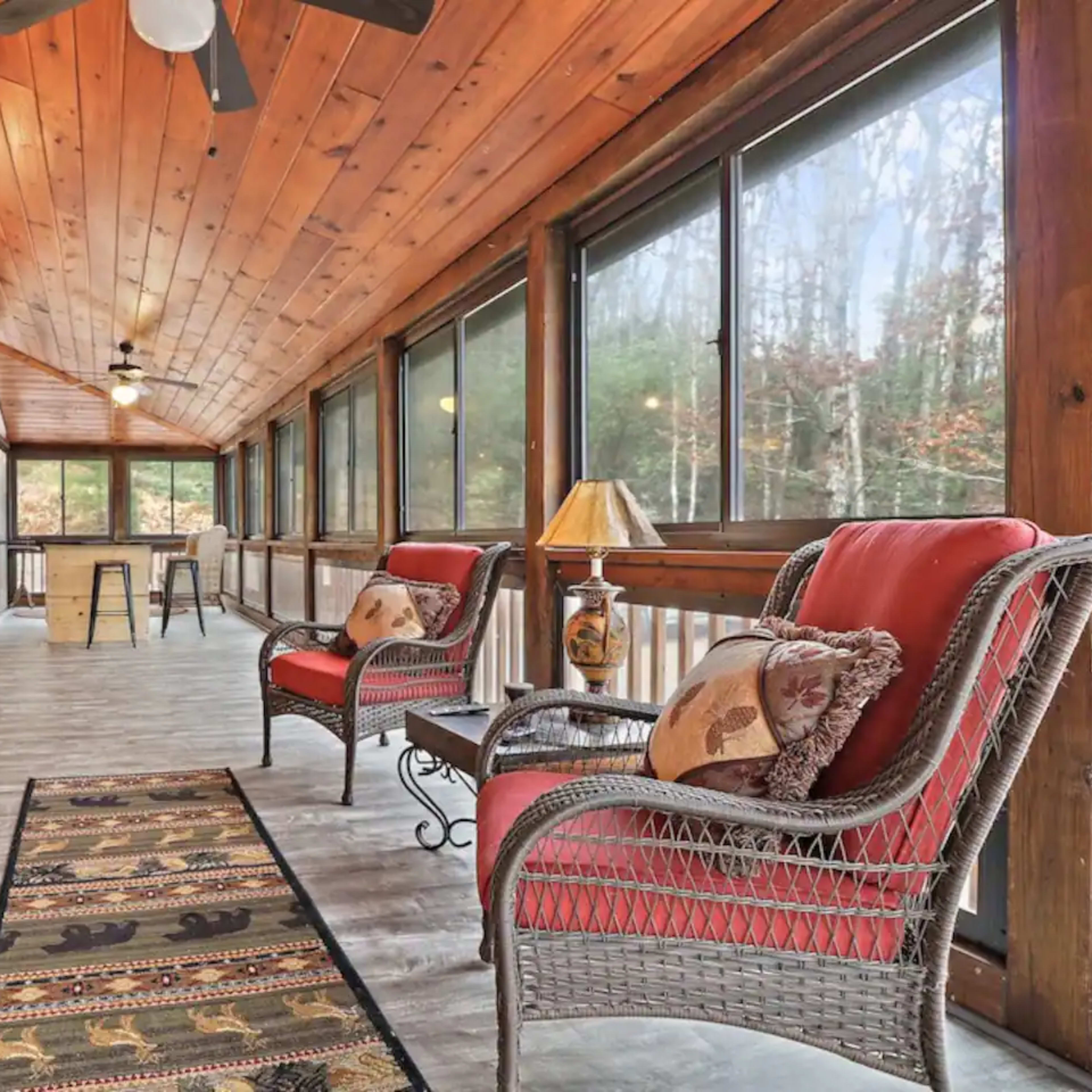 A wooden screened porch with a television, two wicker chairs, a small table, and large windows overlooking a forested area.