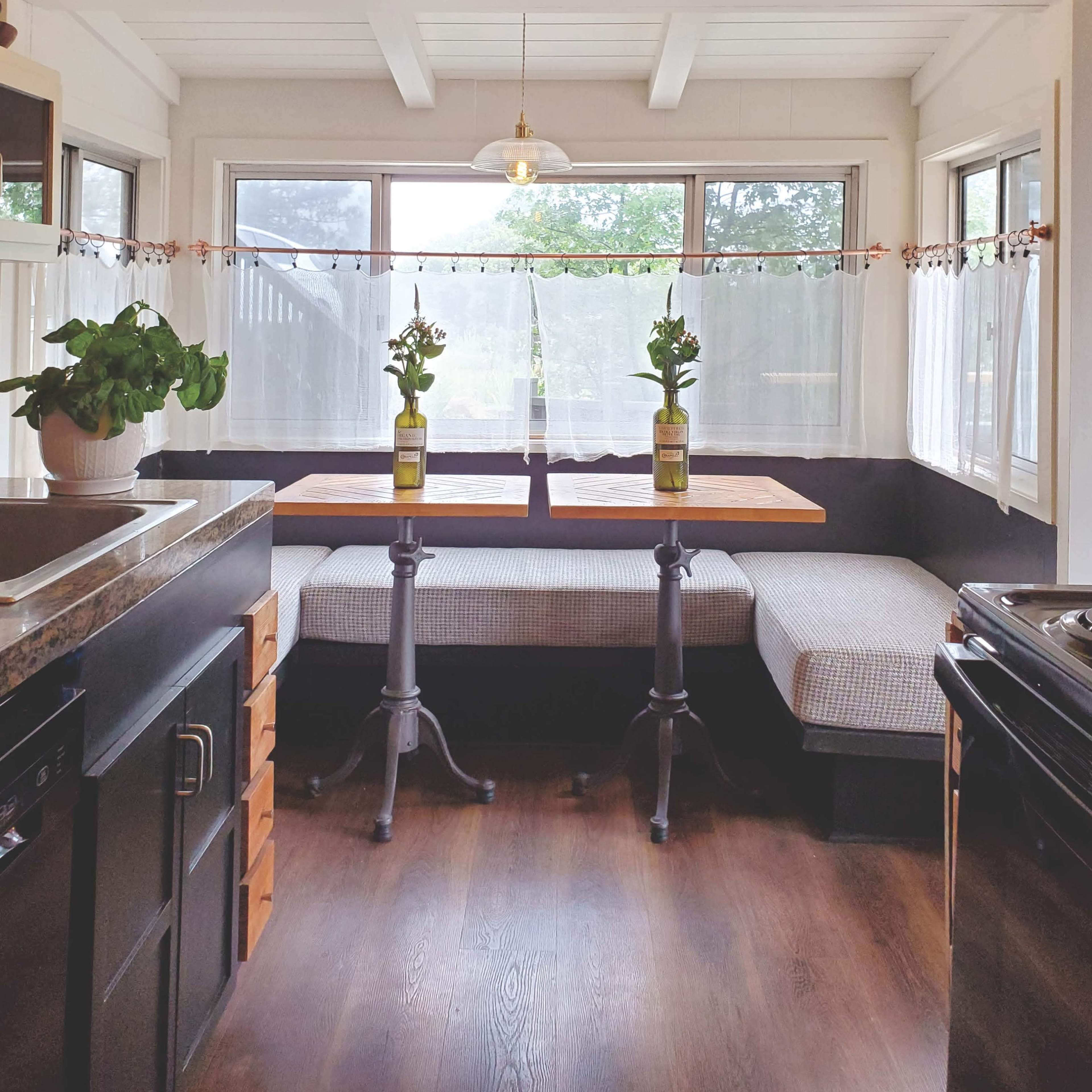 A sunlit dining area features a wooden table with two chairs on a cozy bench, surrounded by large windows and a potted plant.