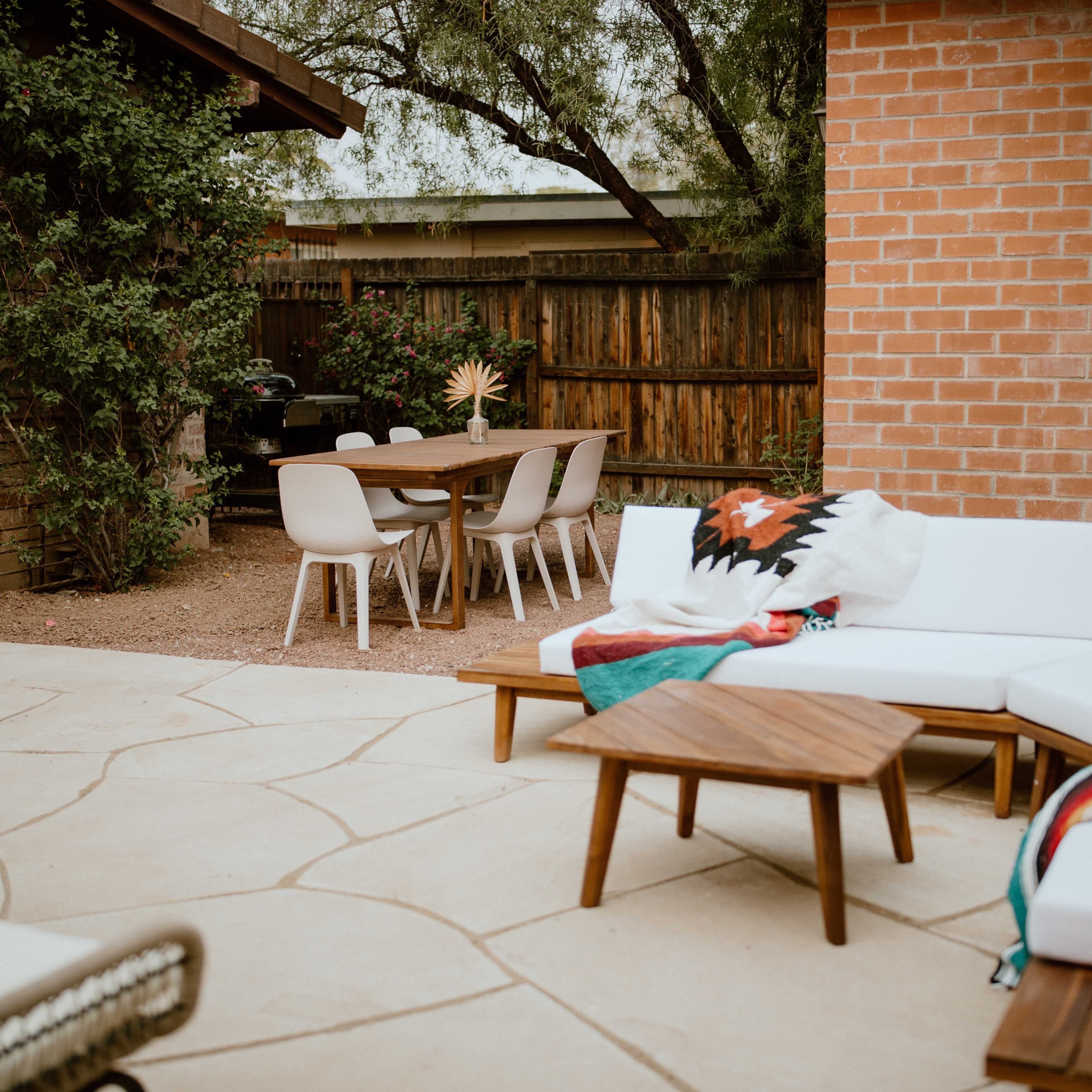 A backyard patio area featuring a sectional sofa with a colorful blanket, a wooden coffee table, and a dining table with chairs under a wooden structure.