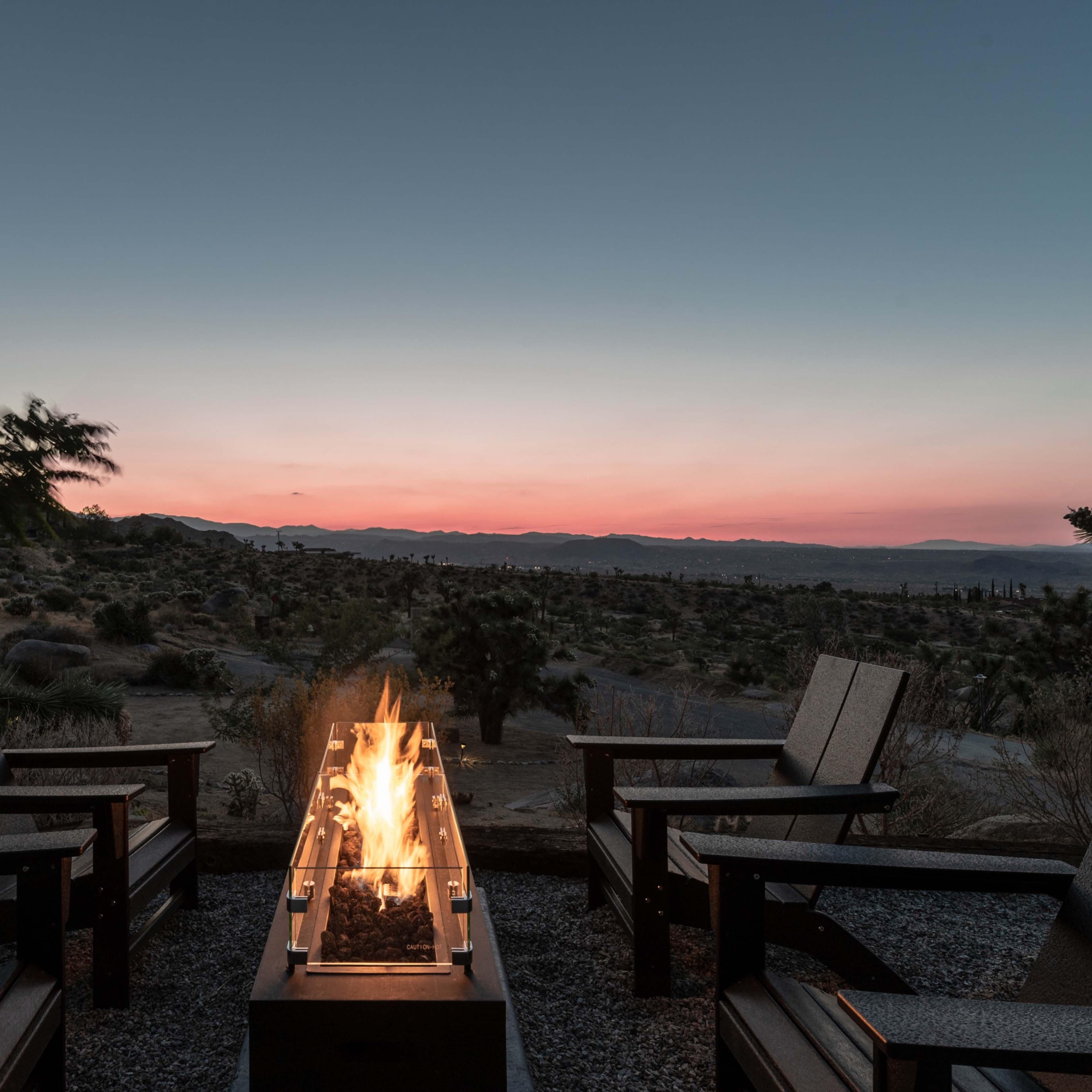 A fire pit with flames is surrounded by four chairs, overlooking a desert landscape at twilight.