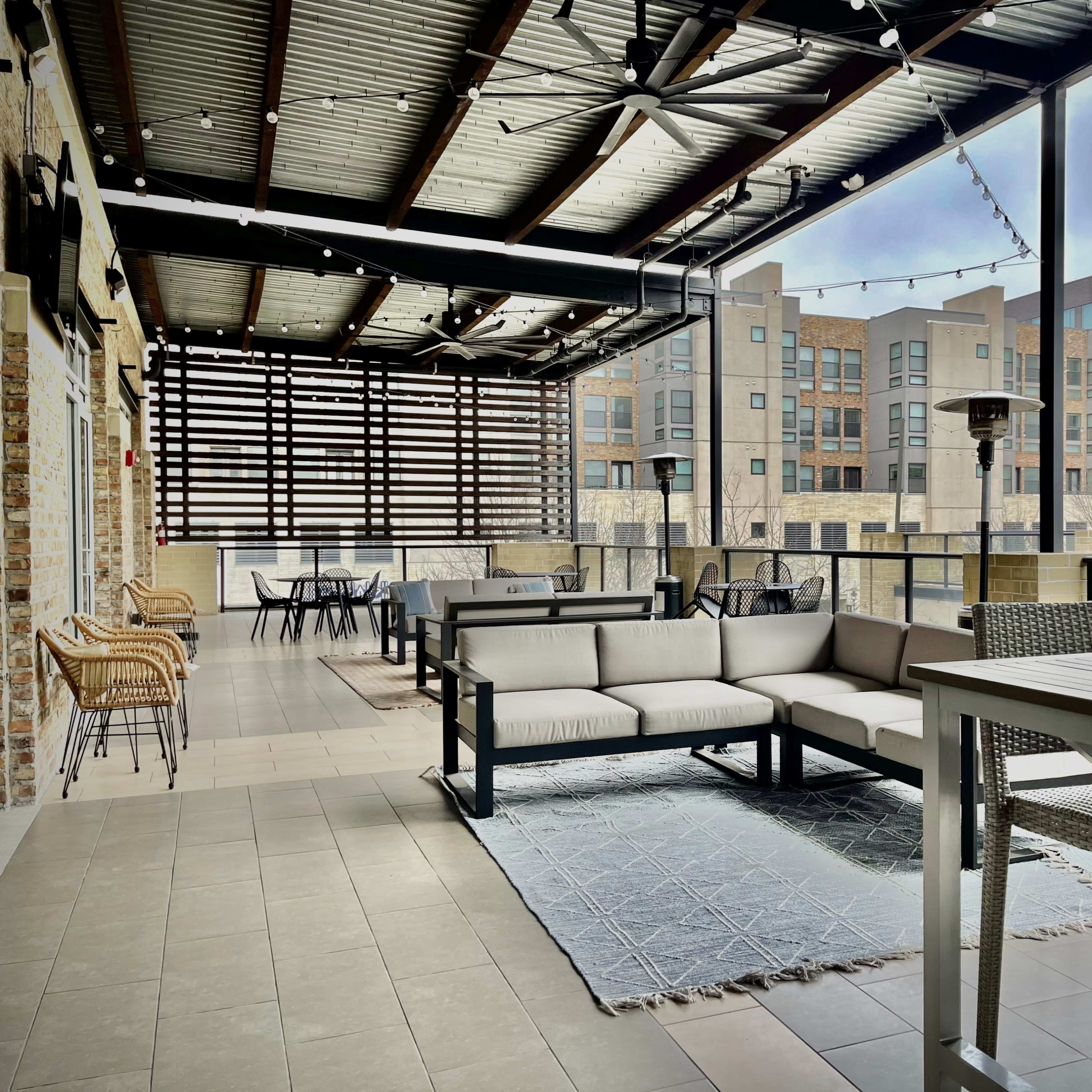 A spacious outdoor terrace with modern seating arrangements, a table, and overhead lighting, overlooking a multi-story building complex.