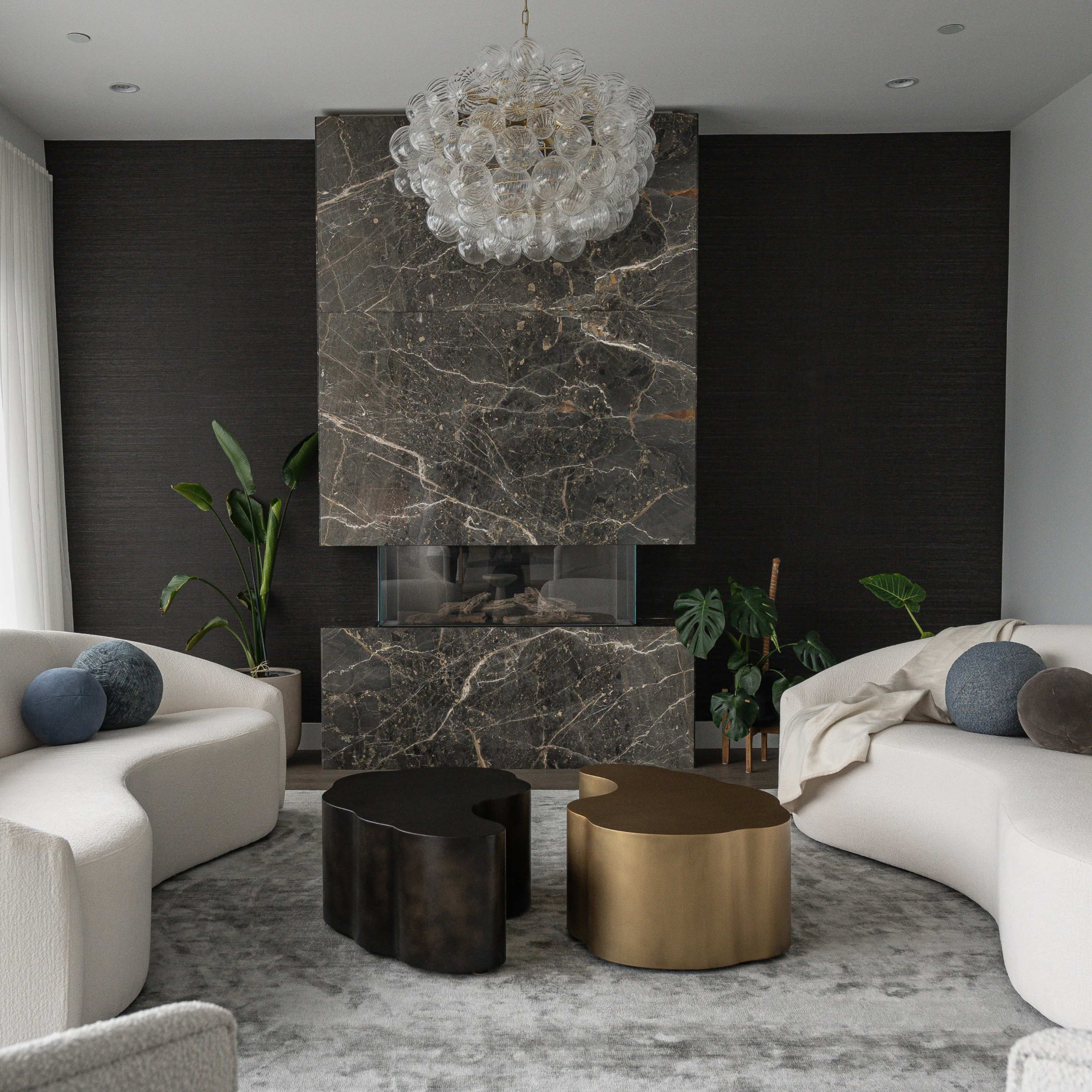 A modern living room features two curved sofas, a marble fireplace with a decorative mantel, and two contemporary coffee tables in dark and metallic finishes.