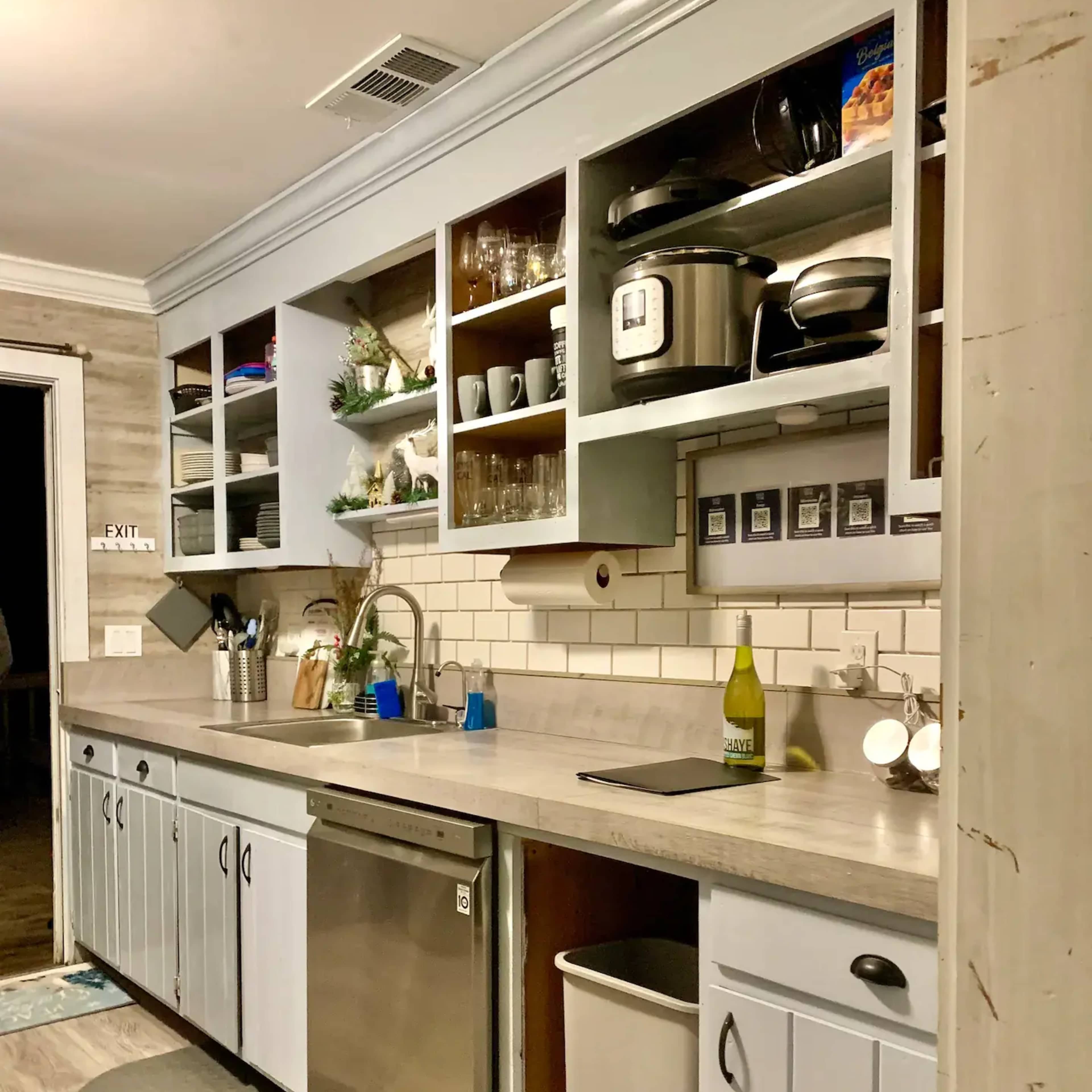 A neatly arranged kitchen features a long countertop, upper shelves with glassware and appliances, a sink, and a dishwasher below.