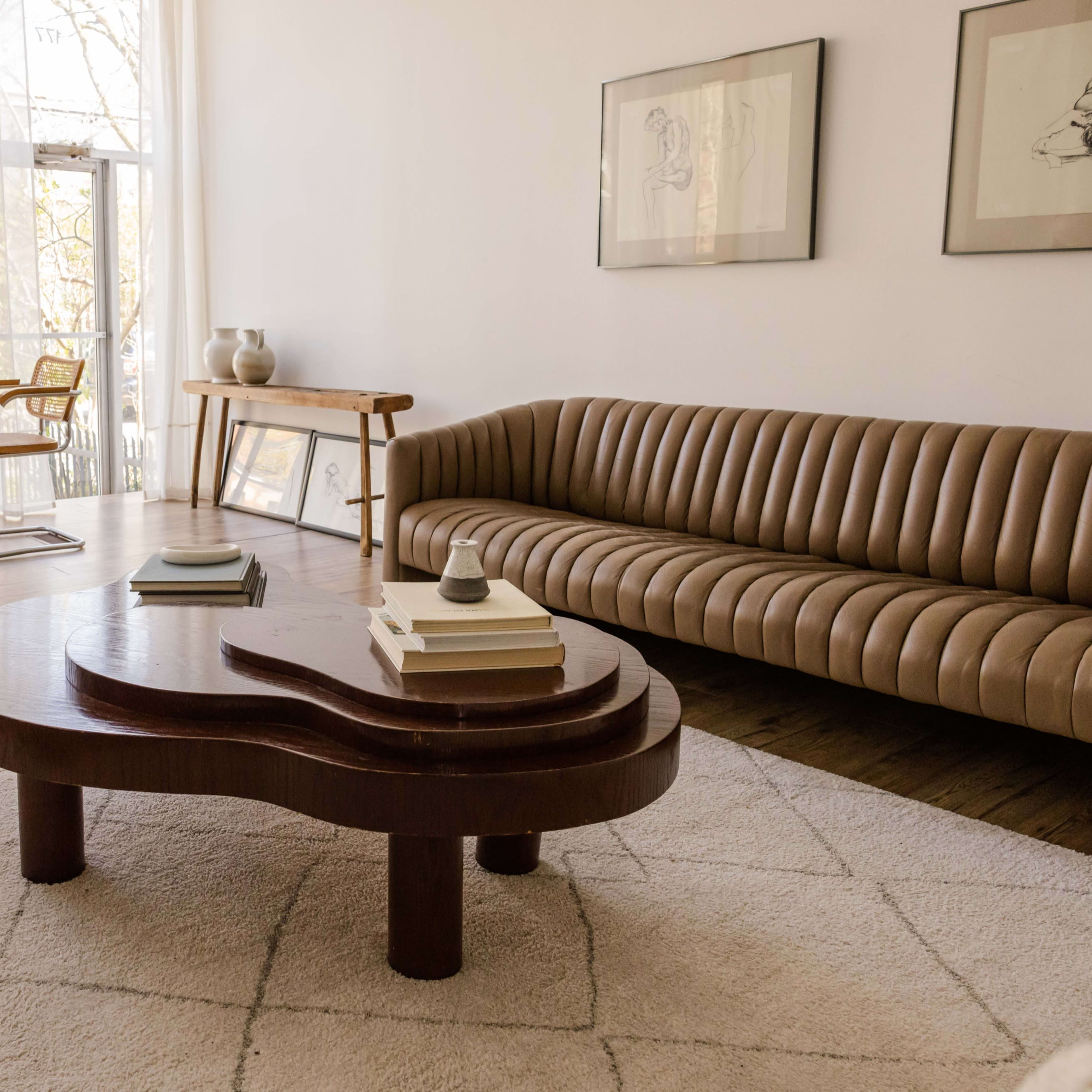 A minimalist living room featuring a curved brown sofa, a wooden coffee table, a patterned rug, and a dining area with chairs and a table.