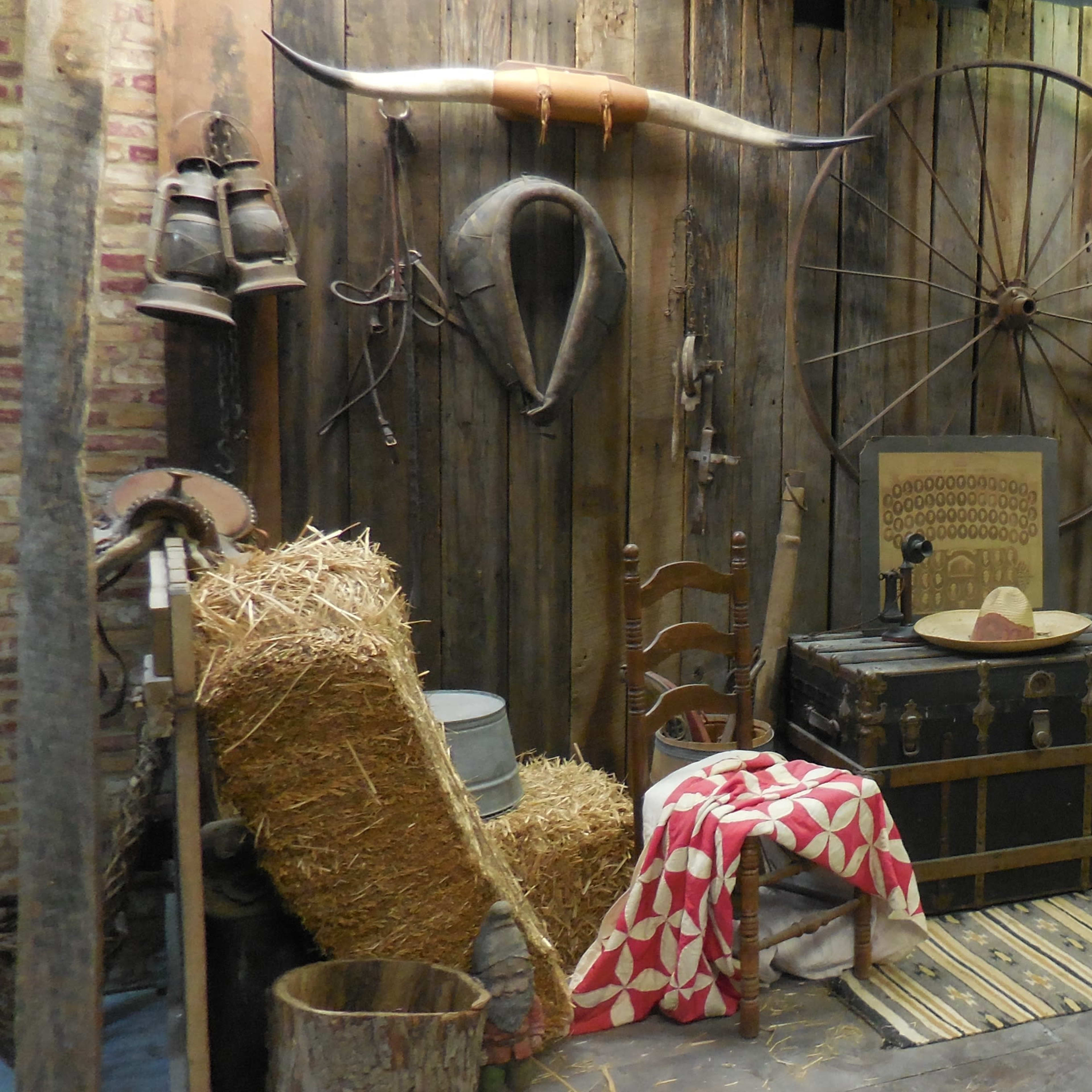 A rustic room decorated with wooden walls, various antique farm tools, a barrel, a chest, and some hay bales.