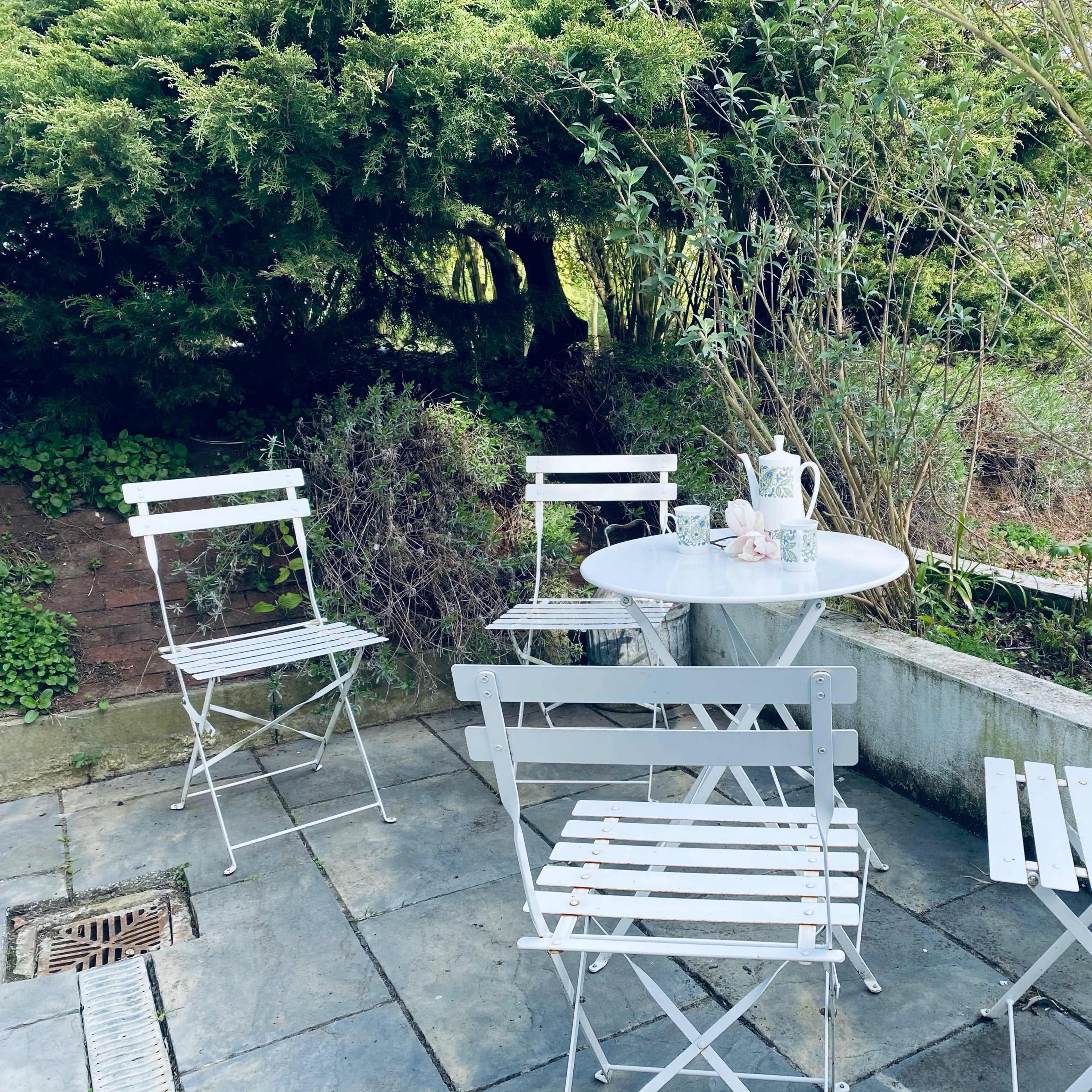 A small outdoor seating area features a round table surrounded by four metal chairs on a stone patio, with greenery in the background.