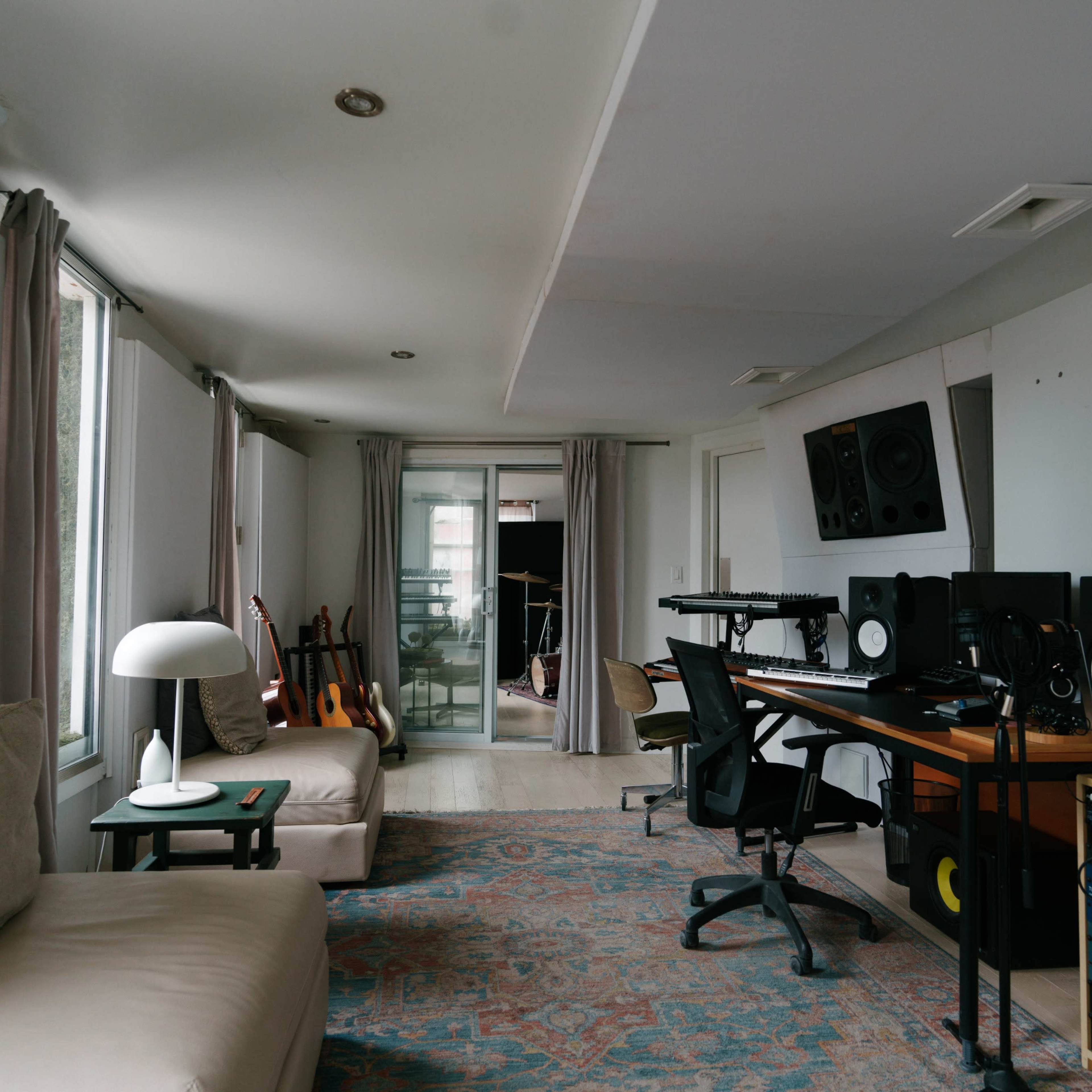 A music studio with a mixing desk, monitors, and instruments, featuring a spacious layout with large windows and modern furnishings.
