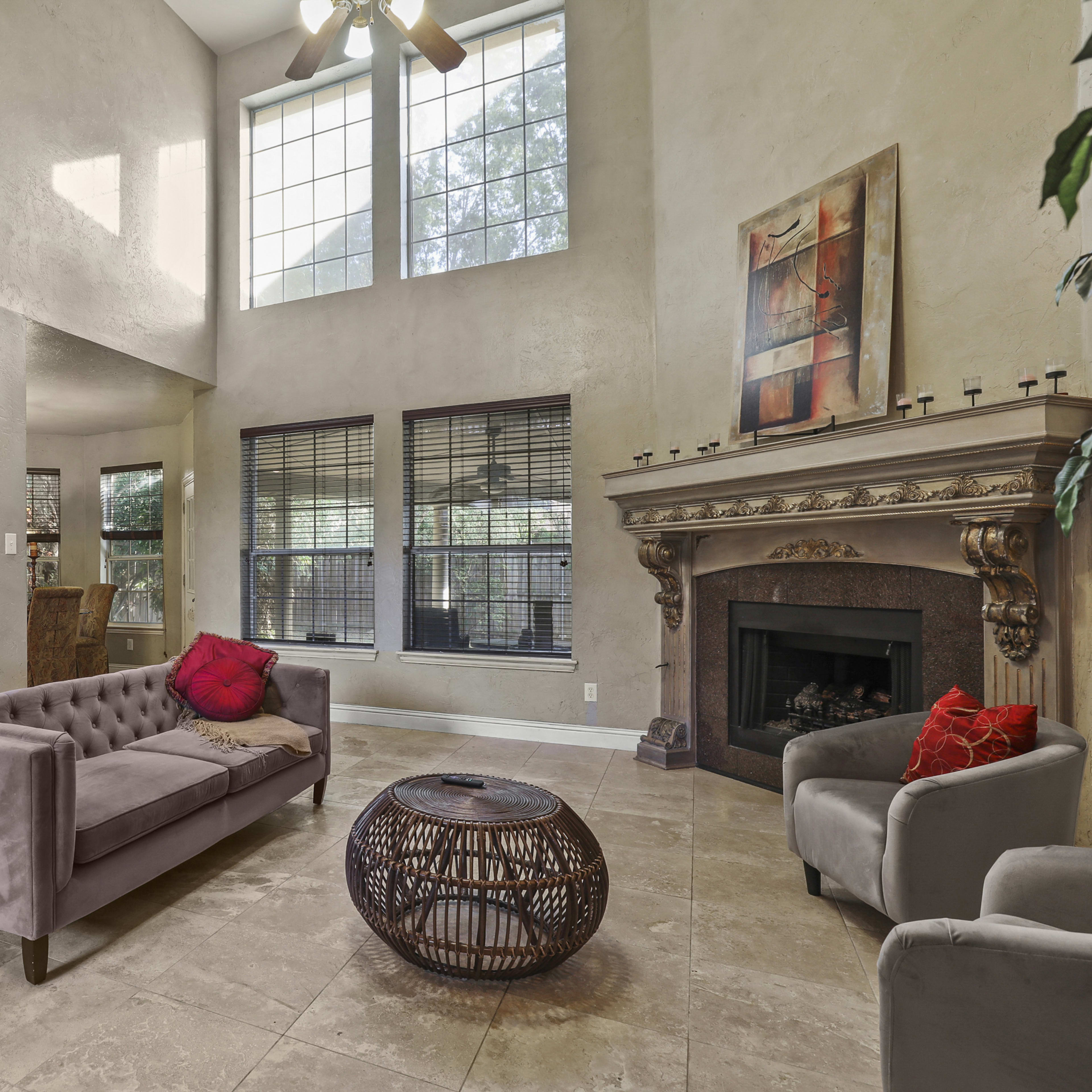 A spacious living area with a high ceiling, featuring a gray tufted sofa, a round wicker coffee table, and a fireplace surrounded by a decorative mantel, with a kitchen area visible in the background.