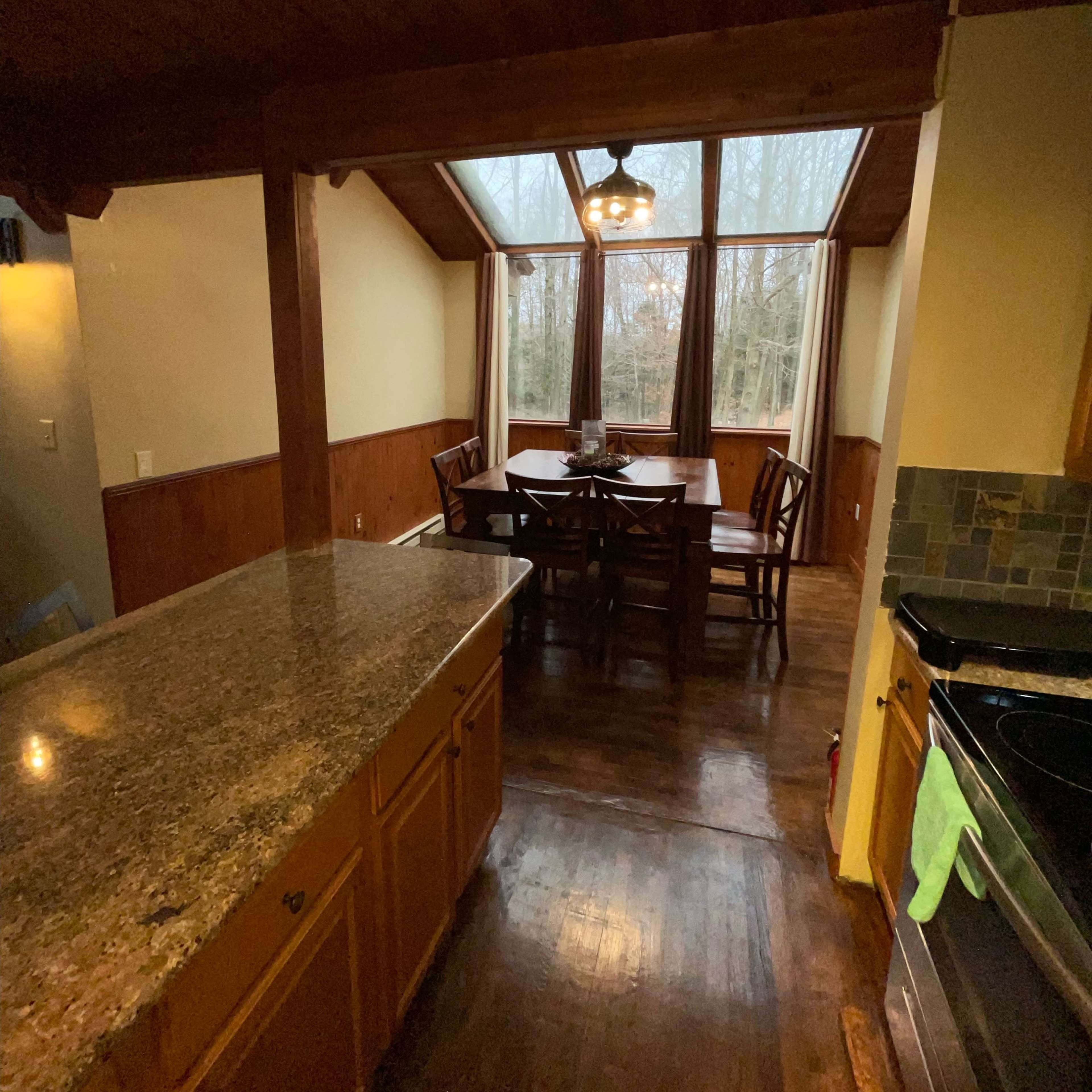 A kitchen with a granite countertop leading into a dining area featuring a large window and a wooden table surrounded by chairs.