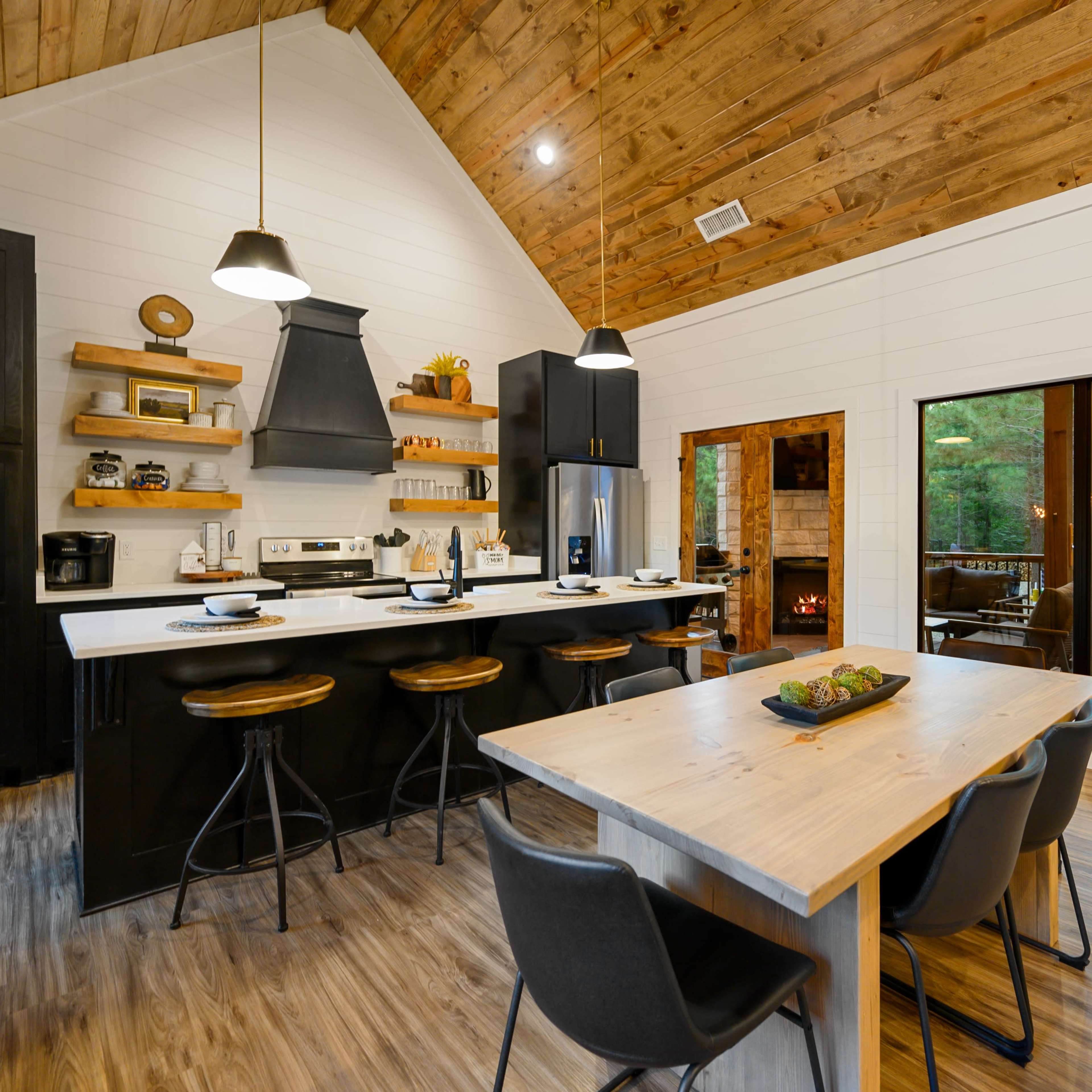 The kitchen features a modern design with black cabinetry, a spacious dining table, and large windows overlooking a wooded area.