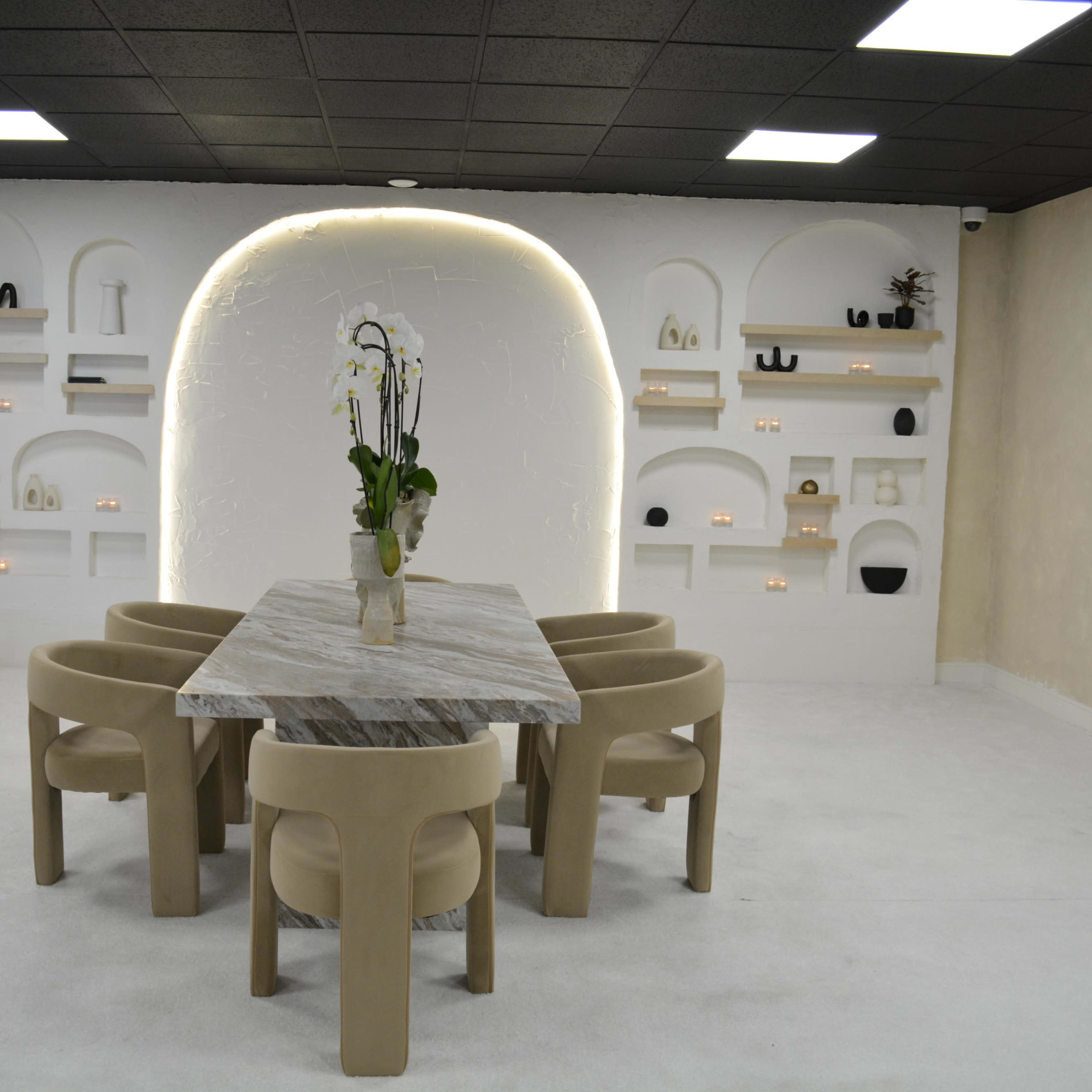 A modern dining space features a marble table surrounded by four beige chairs, with an illuminated arch and a decorative wall shelf in the background.