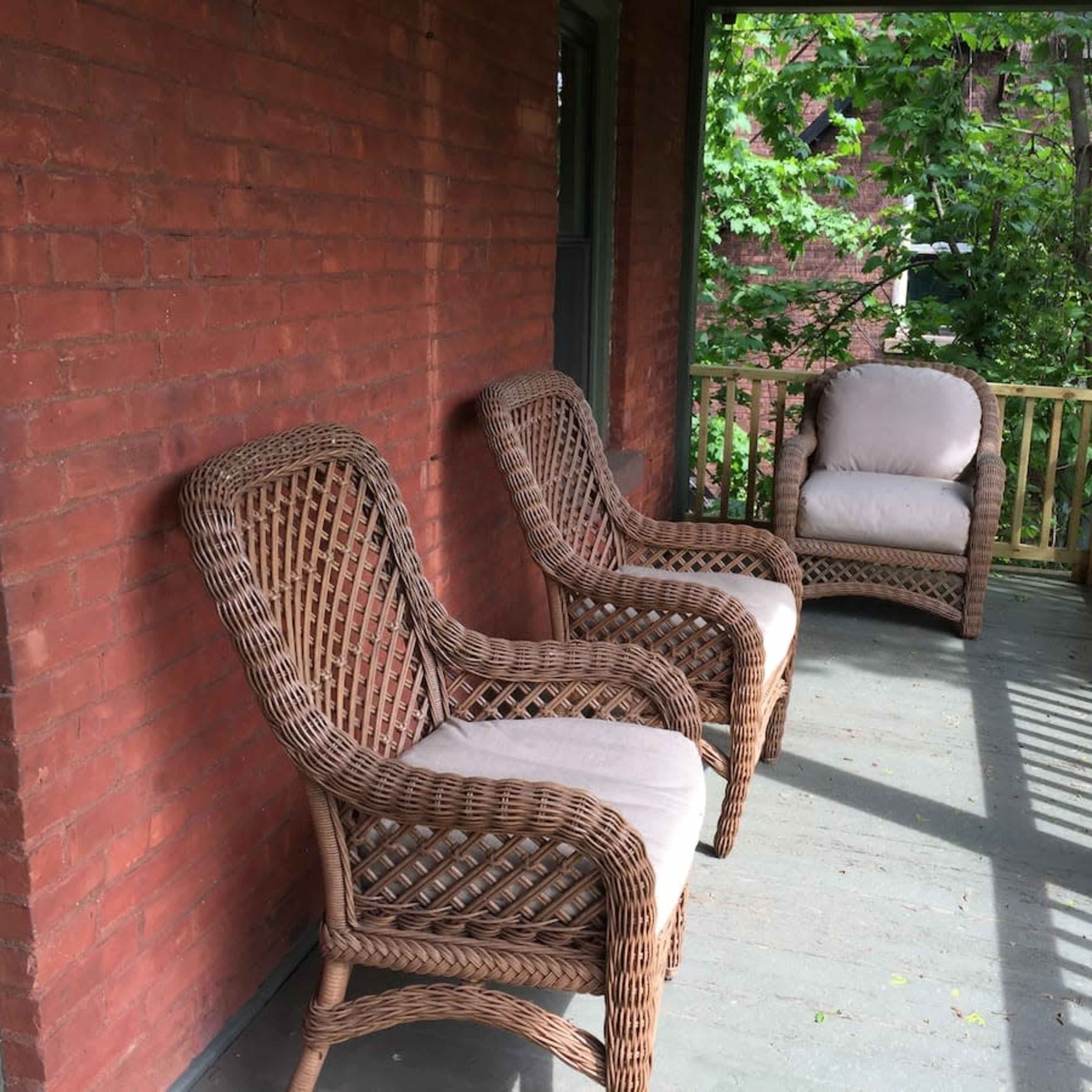 A porch area featuring three wicker chairs positioned on a wood floor, with greenery visible in the background.