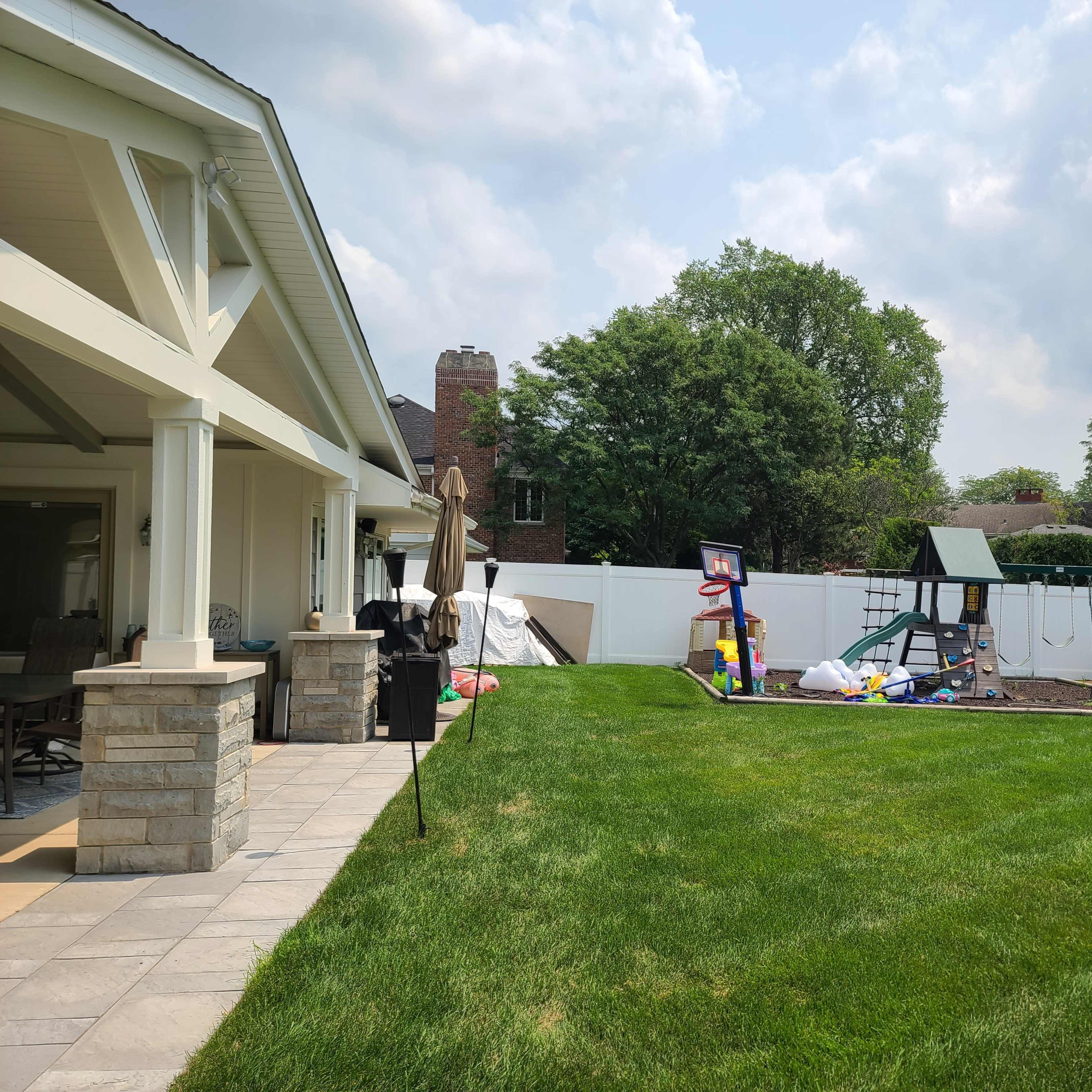 A backyard with a grassy area, a swing set and play structure, and a patio with stone columns adjacent to a house.
