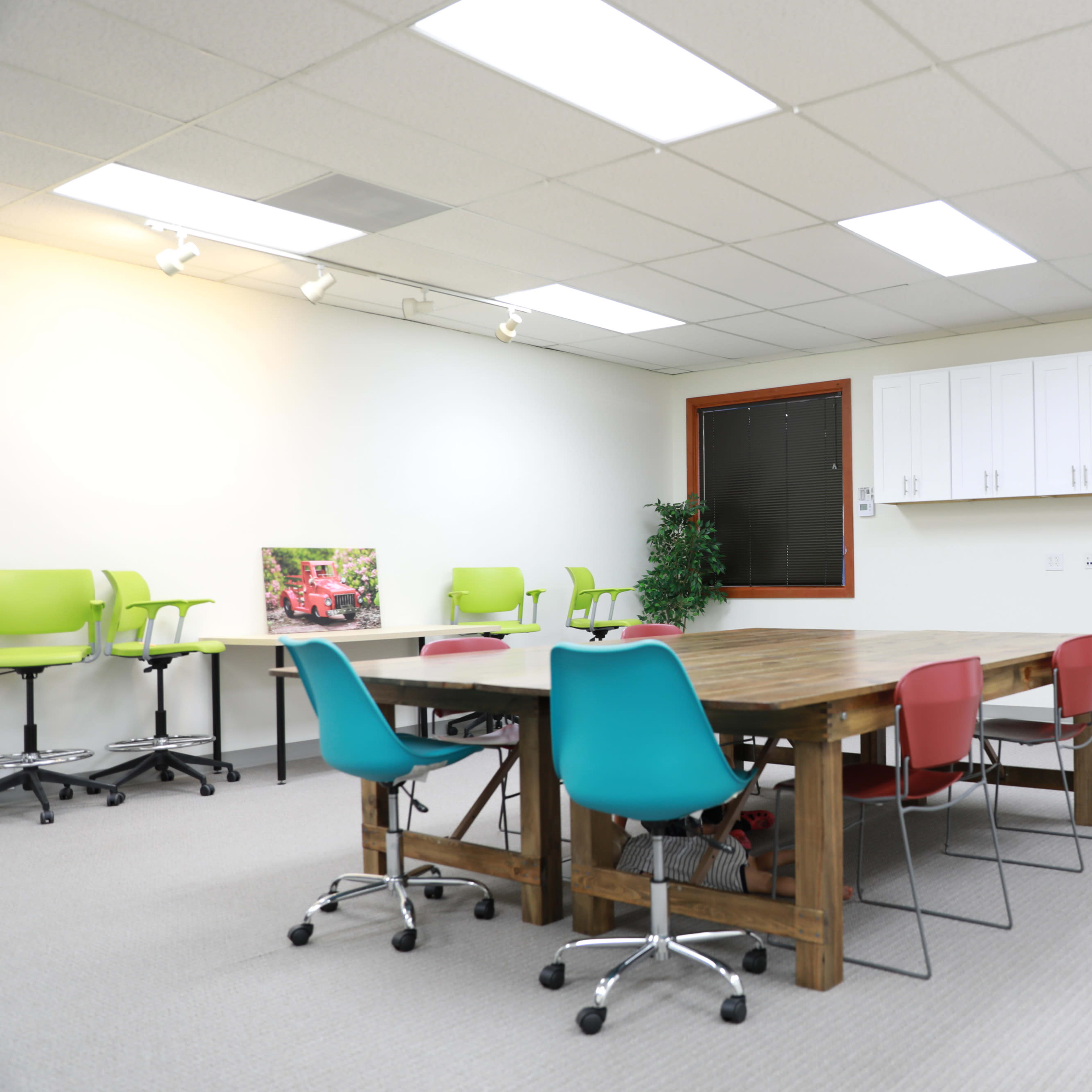 A spacious, well-lit meeting room featuring a large wooden table surrounded by colorful chairs and a few plants.