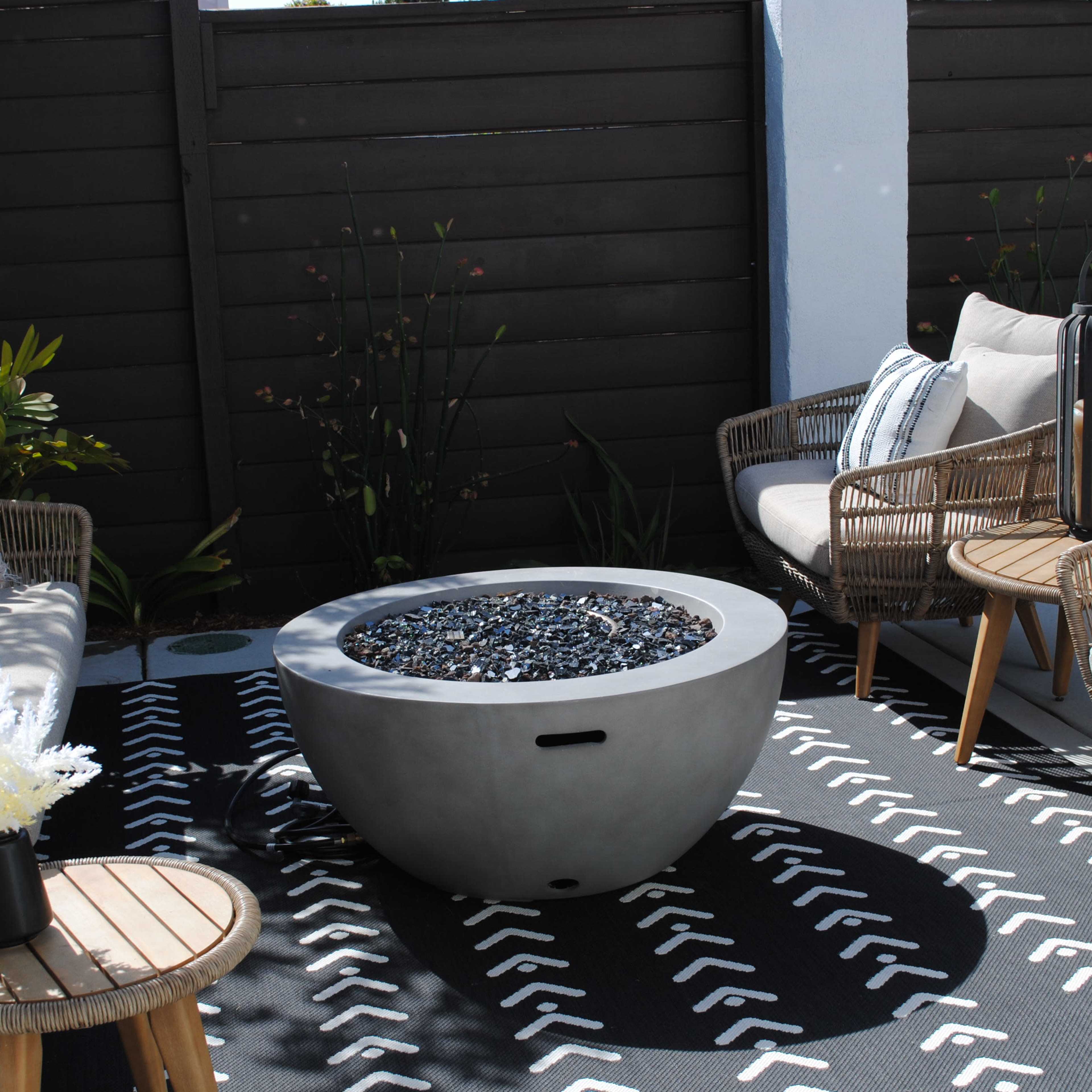 A patio area features a circular concrete fire pit surrounded by four wicker chairs and a small wooden table, with a patterned black and white rug underneath.