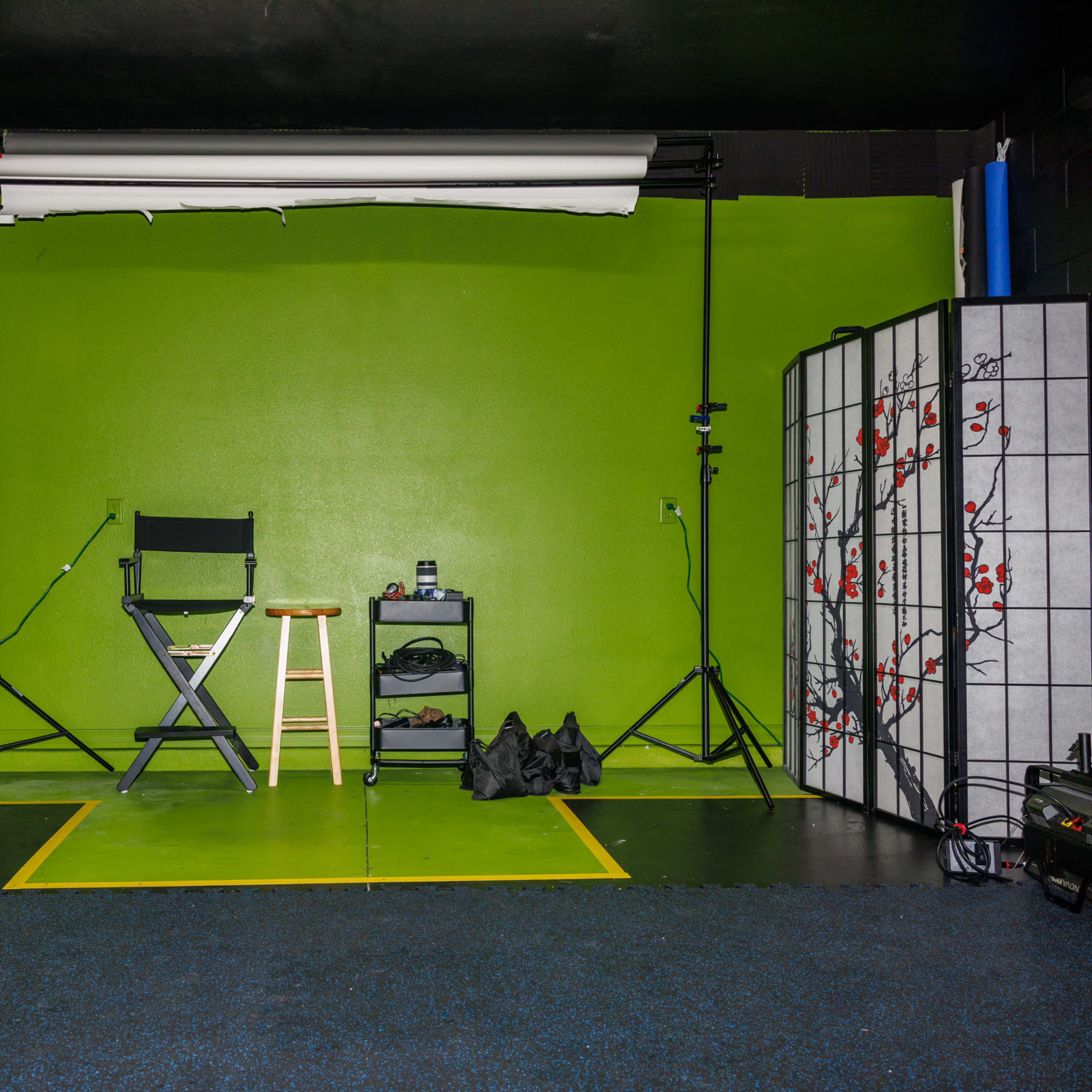 A photography studio with a green backdrop, a director's chair, a stool, lighting equipment, and a black folding screen with a floral design.